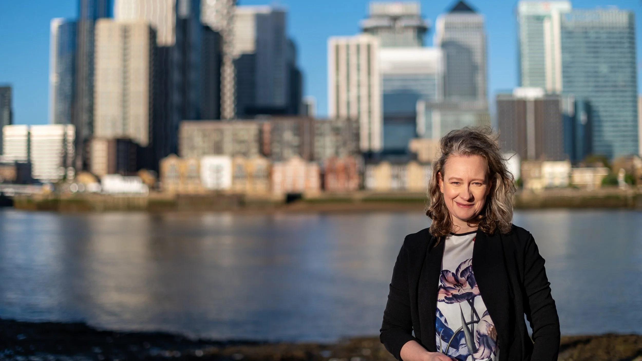 Woman with curly hair standing outdoors near a river with city skyline background