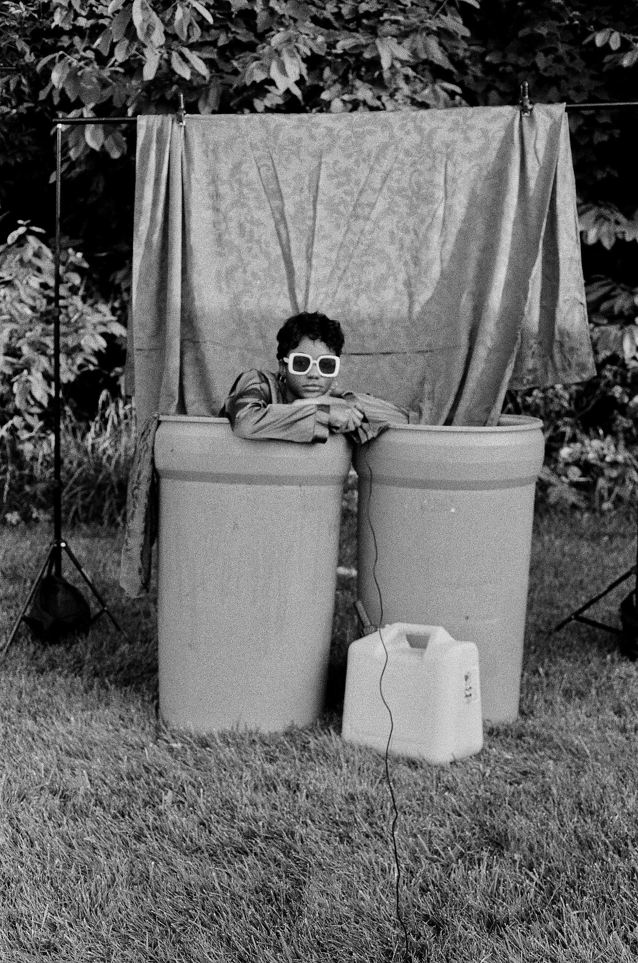Black and white elf Portrait of Christina Rateau holding shutter release cable, inside barrel and perched on the edge of left barrel and looking at the camera. As a backdrop, she uses red fabric draping into two blue barrels.