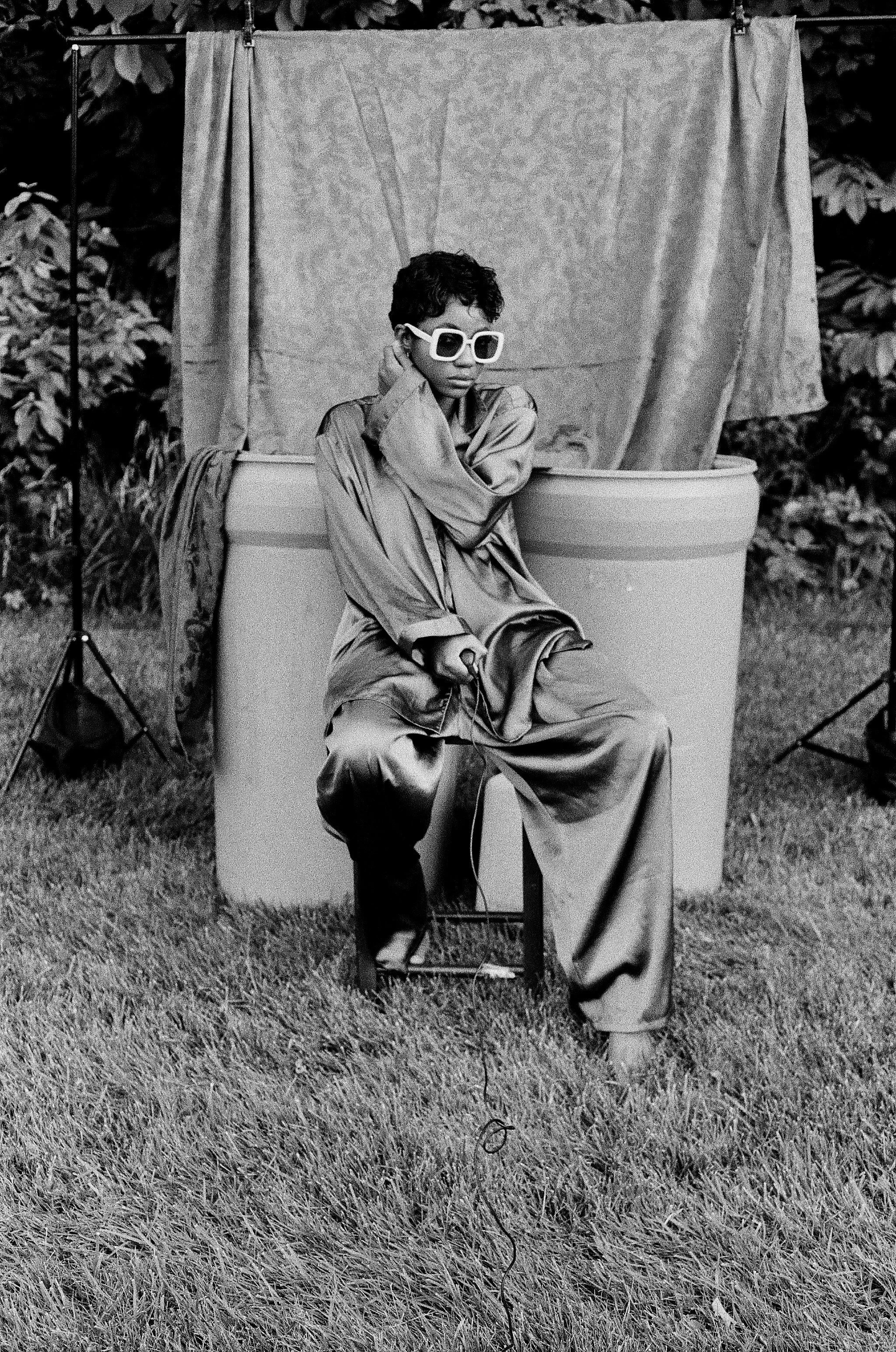 Black and white self portrait of Christina Rateau holding shutter release cable, seated on stool. As a backdrop she uses red fabric draping into two blue barrels with yellow gas can in front of barrels