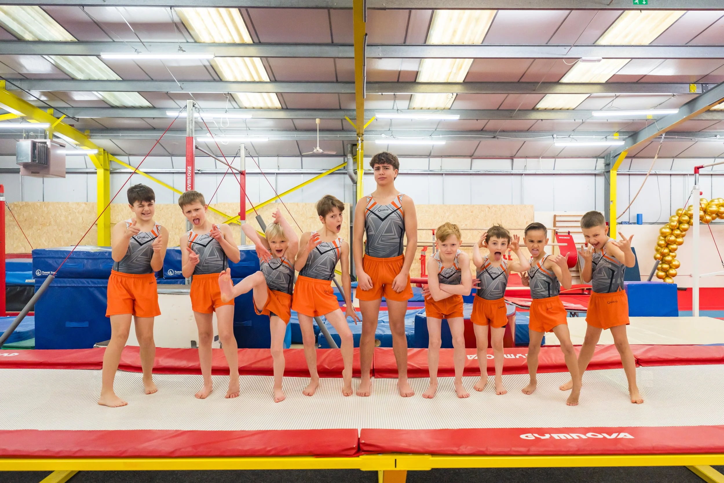 A group of young boys in matching gymnastics uniforms posing on a trampoline inside a gymnastics training facility.