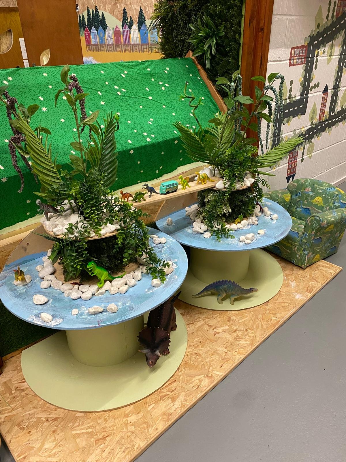 Display with jungle-themed miniature scenery featuring artificial plants, toy dinosaurs, and a toy bus, set on two circular platforms with rocks and water-like surfaces, against a background with colorful paintings and a wall with cityscape designs.
