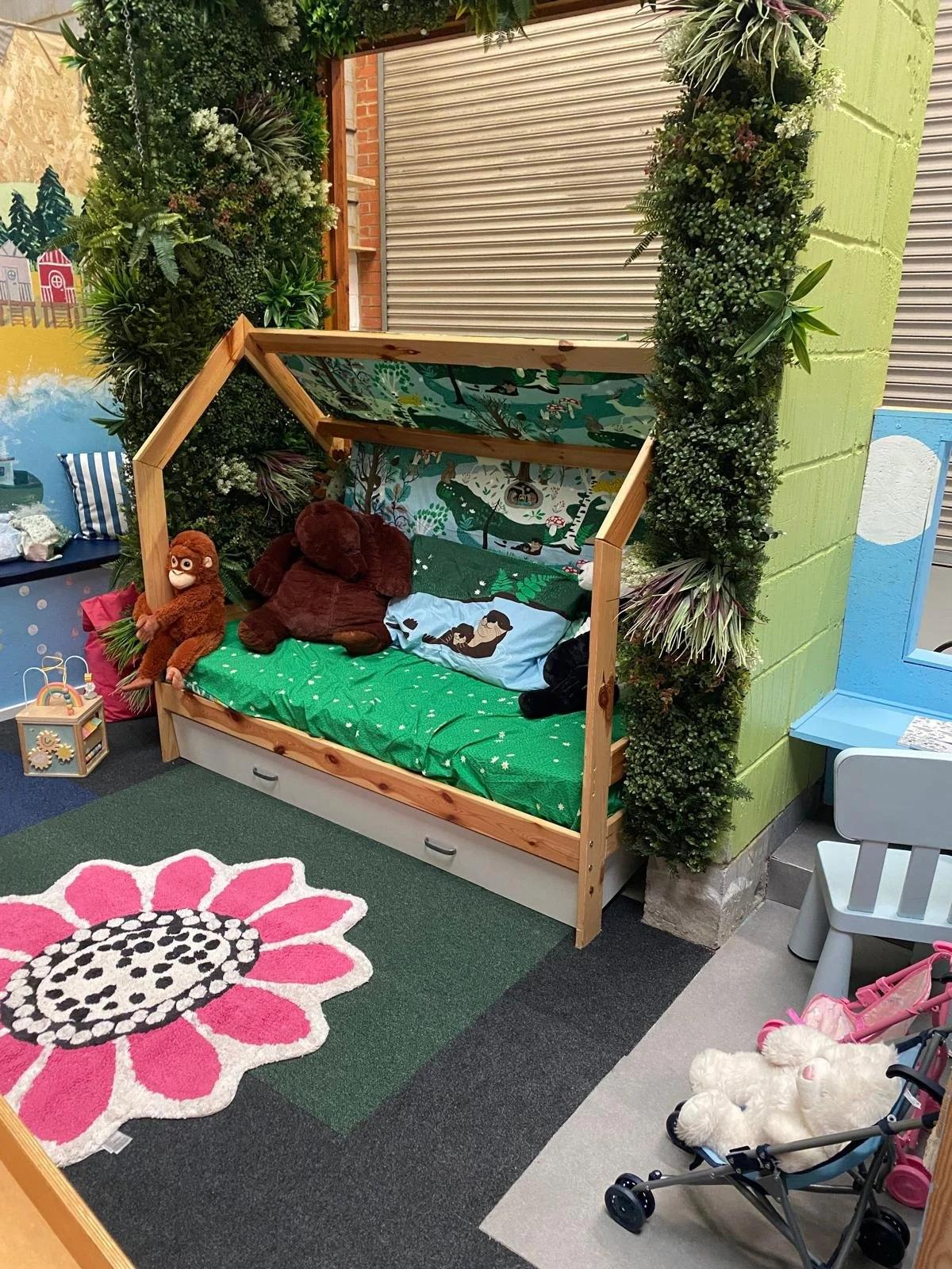 A children's play area with a house-shaped bed surrounded by artificial greenery and toys, including stuffed animals, a stroller with a plush toy, and a colorful flower rug.