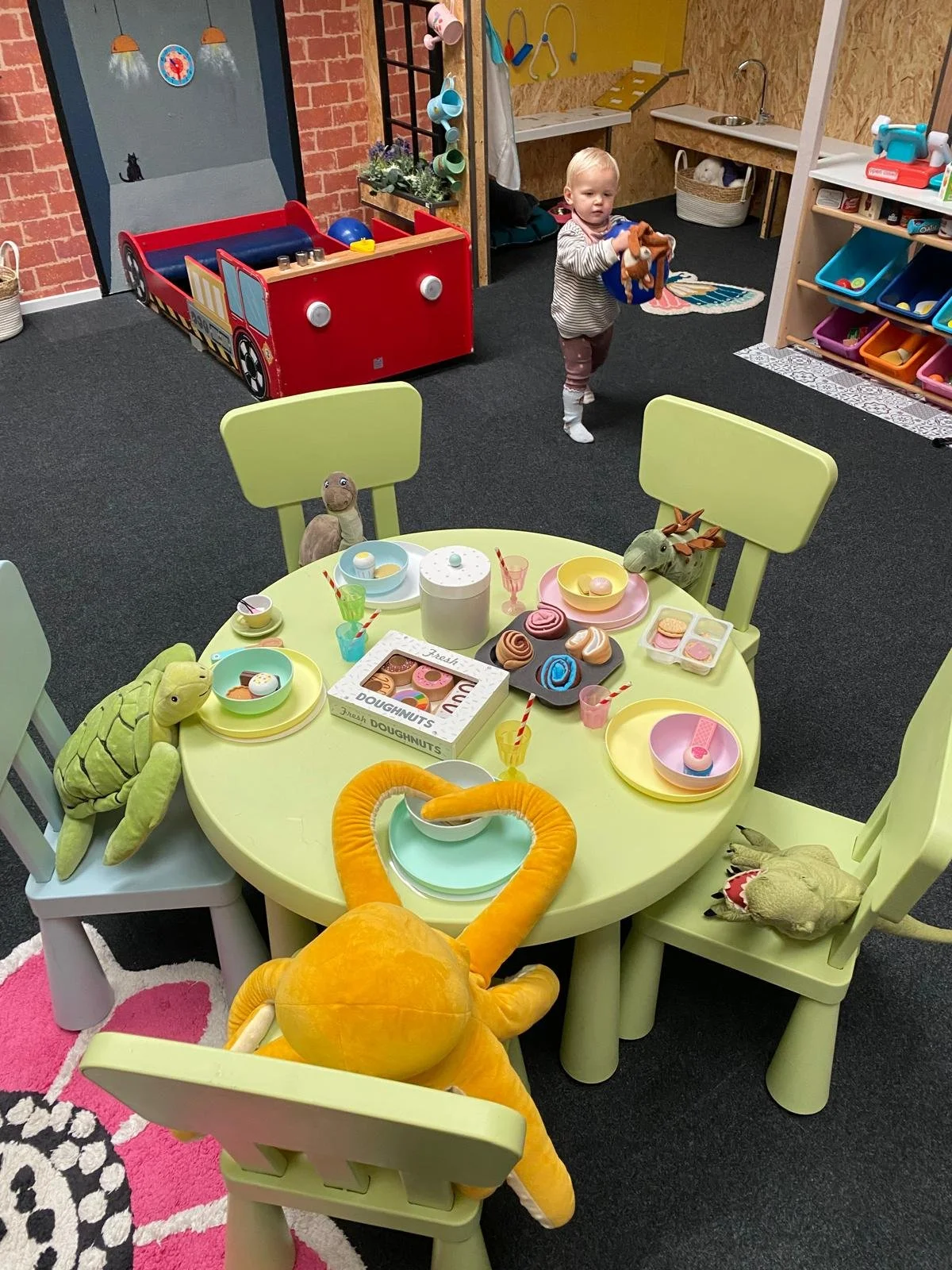 A playroom with a round table set up for pretend tea, featuring plush toys including a turtle, a dragon, and a snake. The table has toy food, plates, cups, and a box of toy donuts. A young child is standing in the background holding a toy, with a play kitchen and storage shelves nearby.
