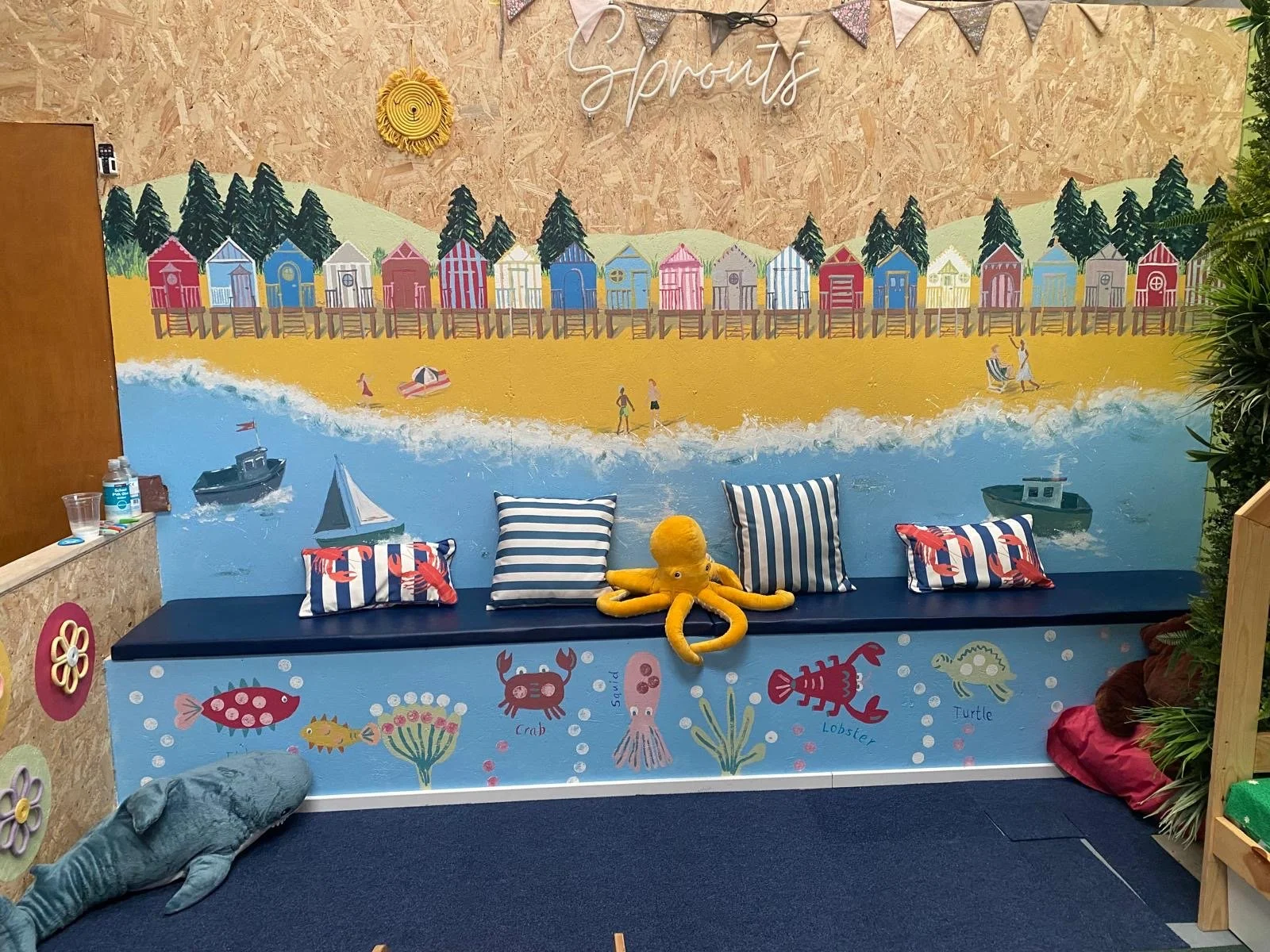 Colorful beach scene mural with beach houses, pine trees, ocean, boats, and people, labeled 'Sprouts'.