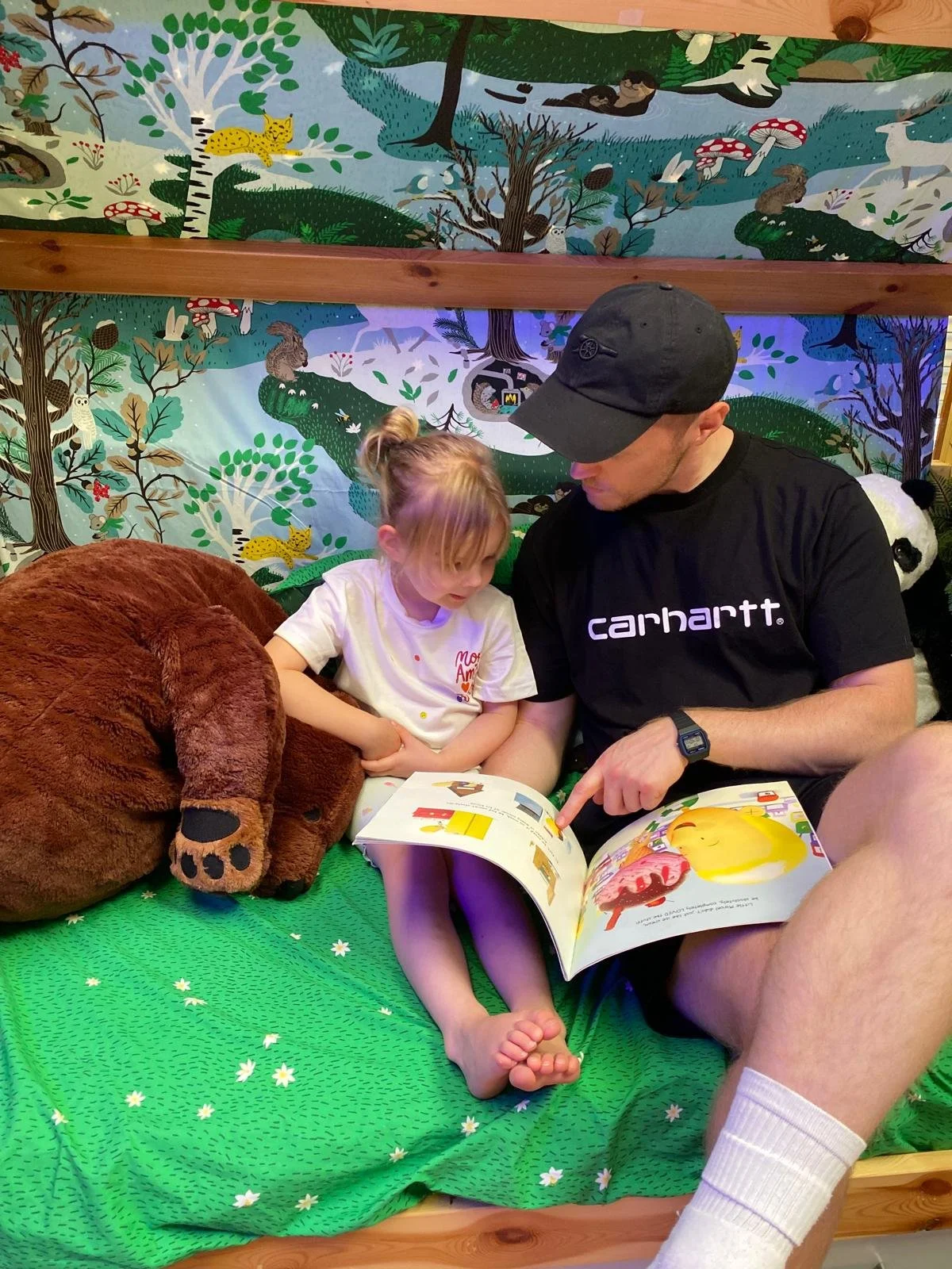A man and a young girl sit together on a green bedspread with daisies, reading a children's book. A large brown stuffed animal is beside them, and a colorful woodland-themed mural decorates the background wall.