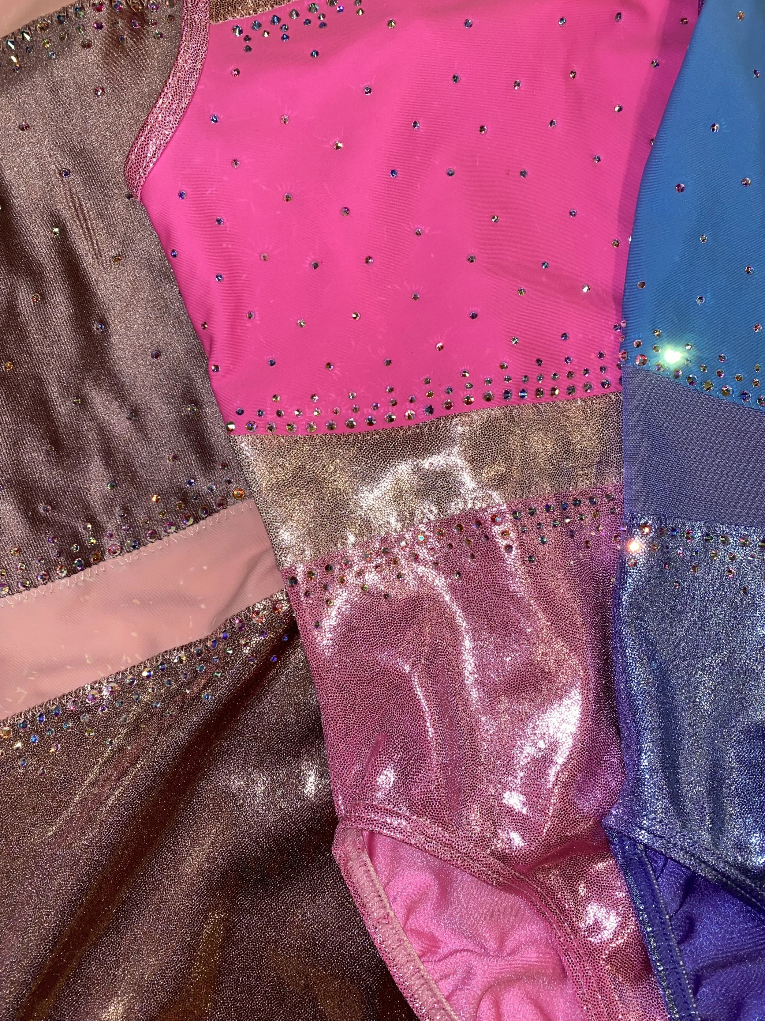 Close-up view of a colorful fabric material decorated with rhinestones, featuring pink, blue, purple, and metallic sections with shiny and glittery textures.