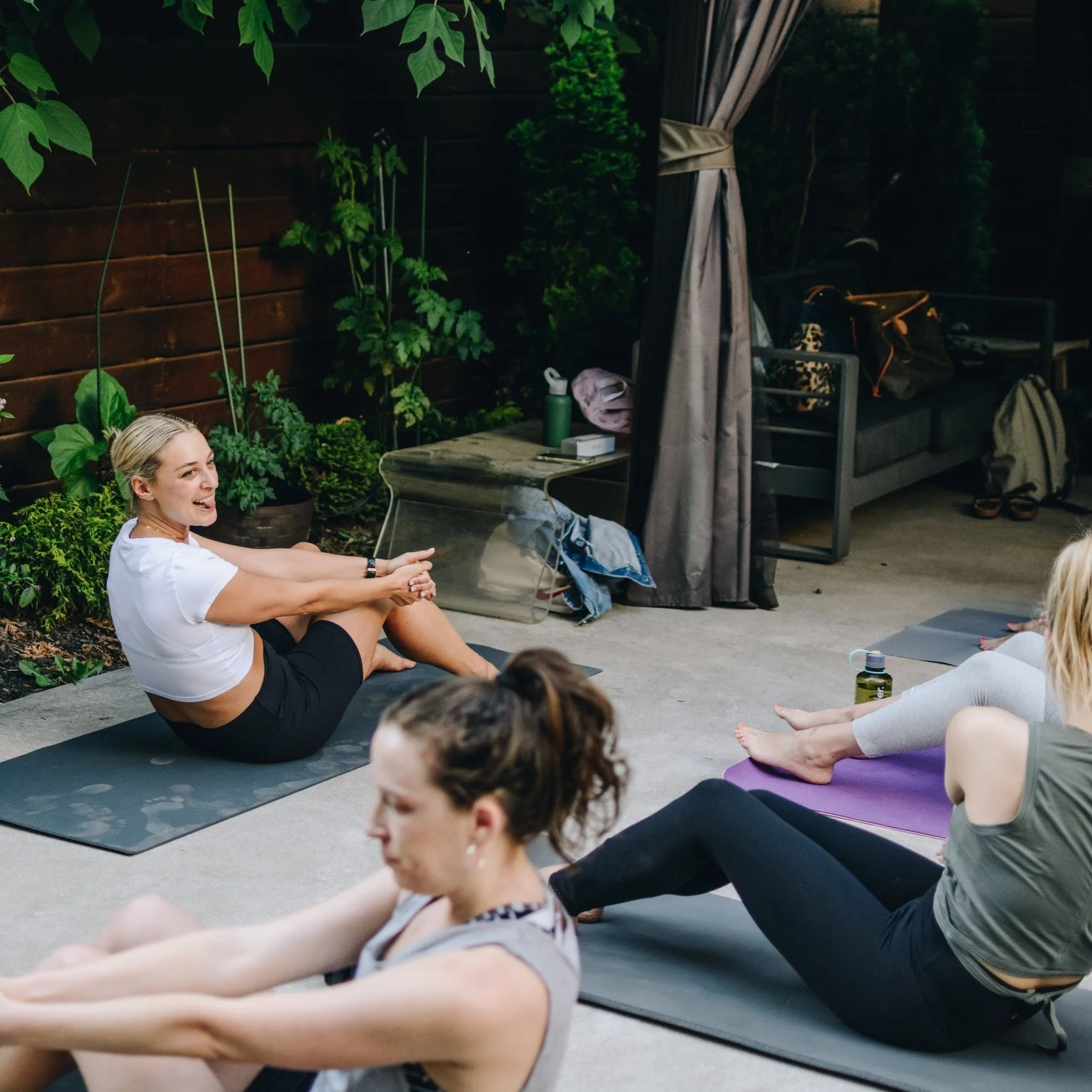 Women participating in an outdoor Pilates with Abs class on mats in a patio with greenery and a wooden fence.