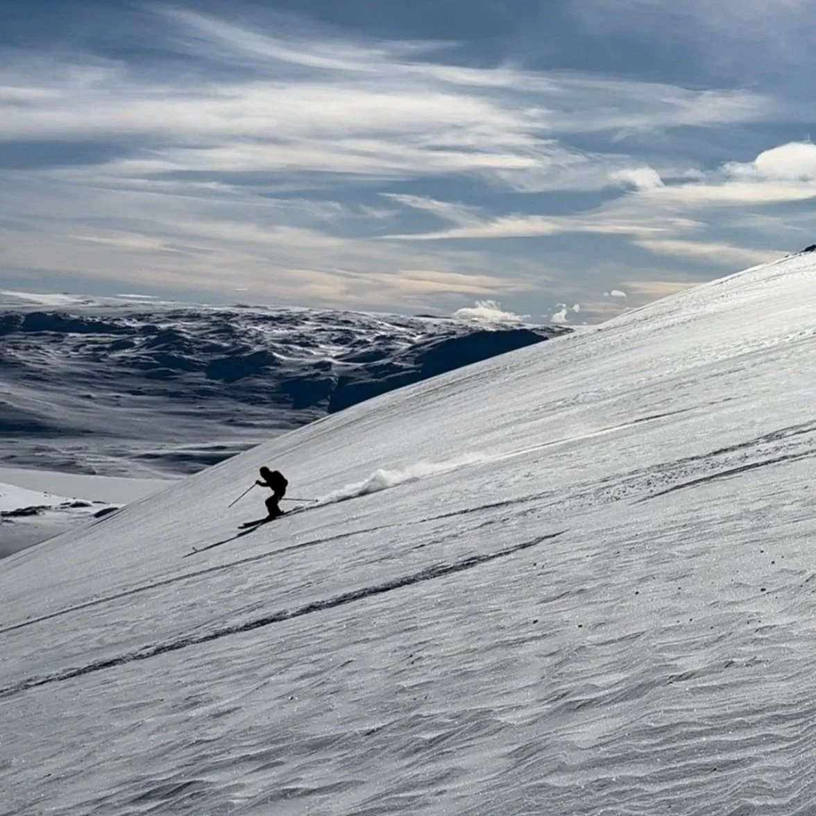 En person som kjører ned en slak fjellside på ski. I bakgrunnen ser man fjell og blå himmel med noen skyer