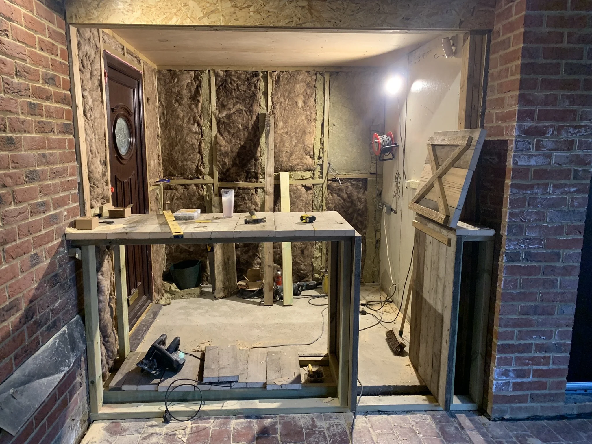 Construction site with a partially built wooden structure, insulation visible on walls, tools like a circular saw and a level on a plywood work surface, and a brick wall backdrop.