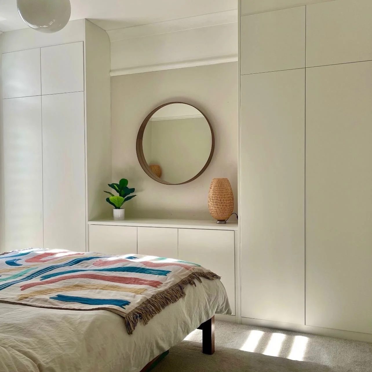Minimalist bedroom with white cabinets, a bed with a colorful blanket, a round mirror, a potted plant, and a wicker lamp on a dresser.