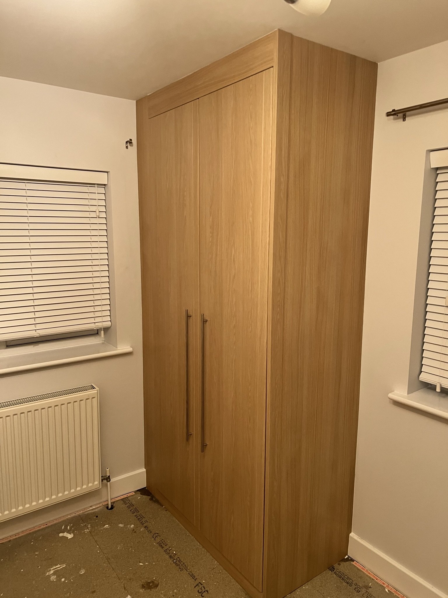 A wooden wardrobe with two tall handles beside a window with blinds in a room. The floor appears unfinished, revealing a concrete surface. A radiator is installed beneath the window.