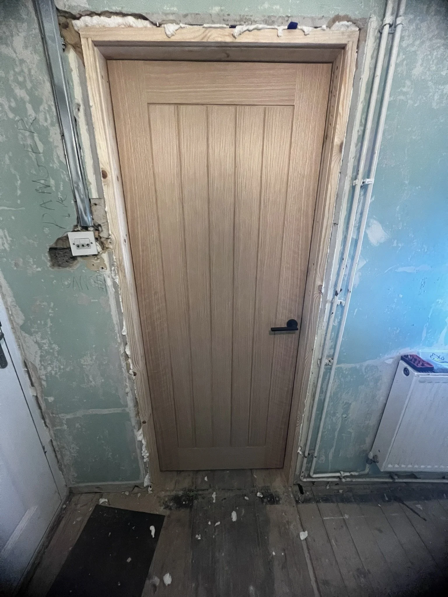 Unfinished interior door installation in a room with exposed drywall and pipes, a wooden door with a black handle, and visible construction debris on the floor.
