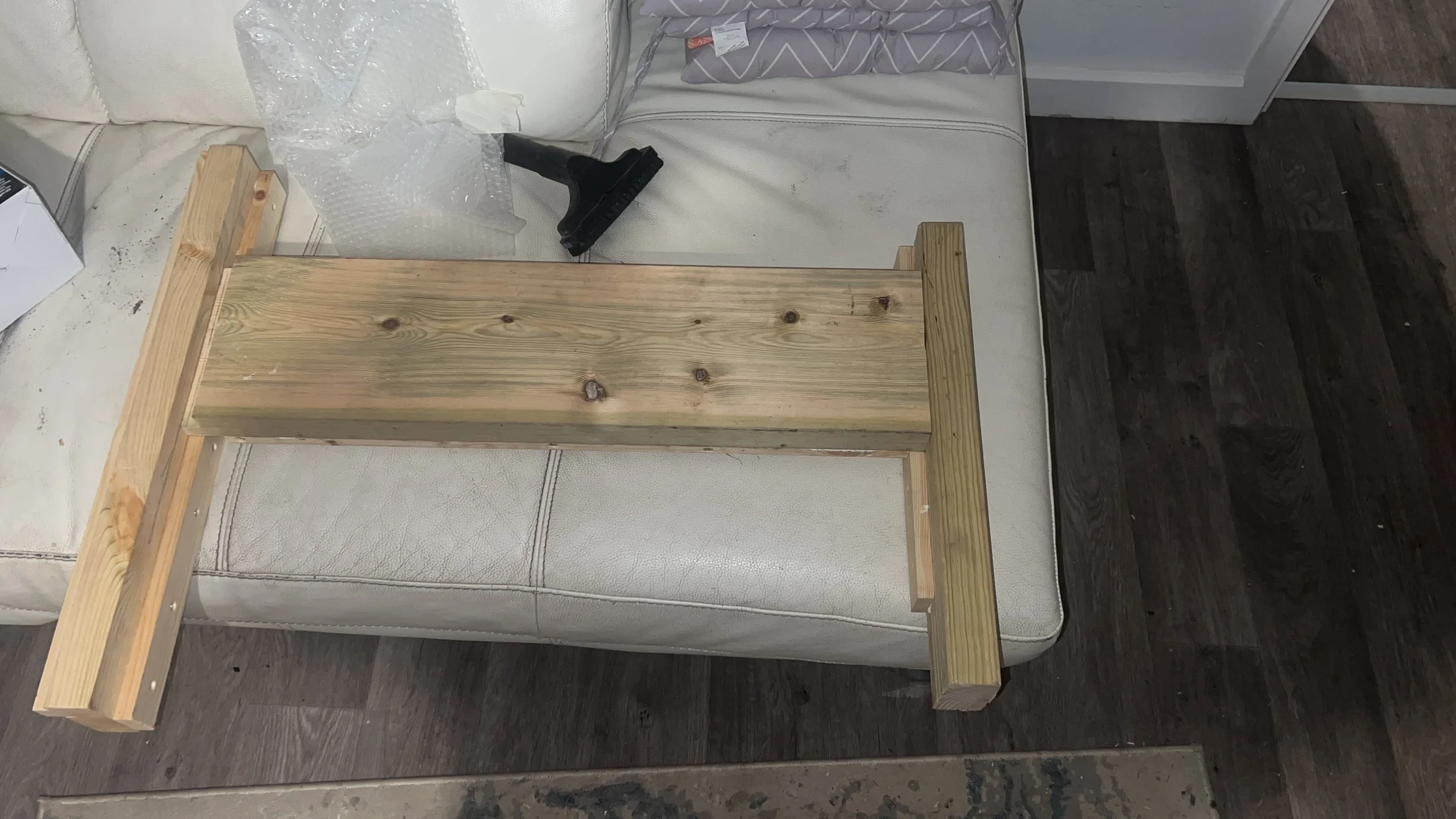 Partially assembled wooden table on white couch with bubble wrap, vacuum attachment, and cushions nearby.