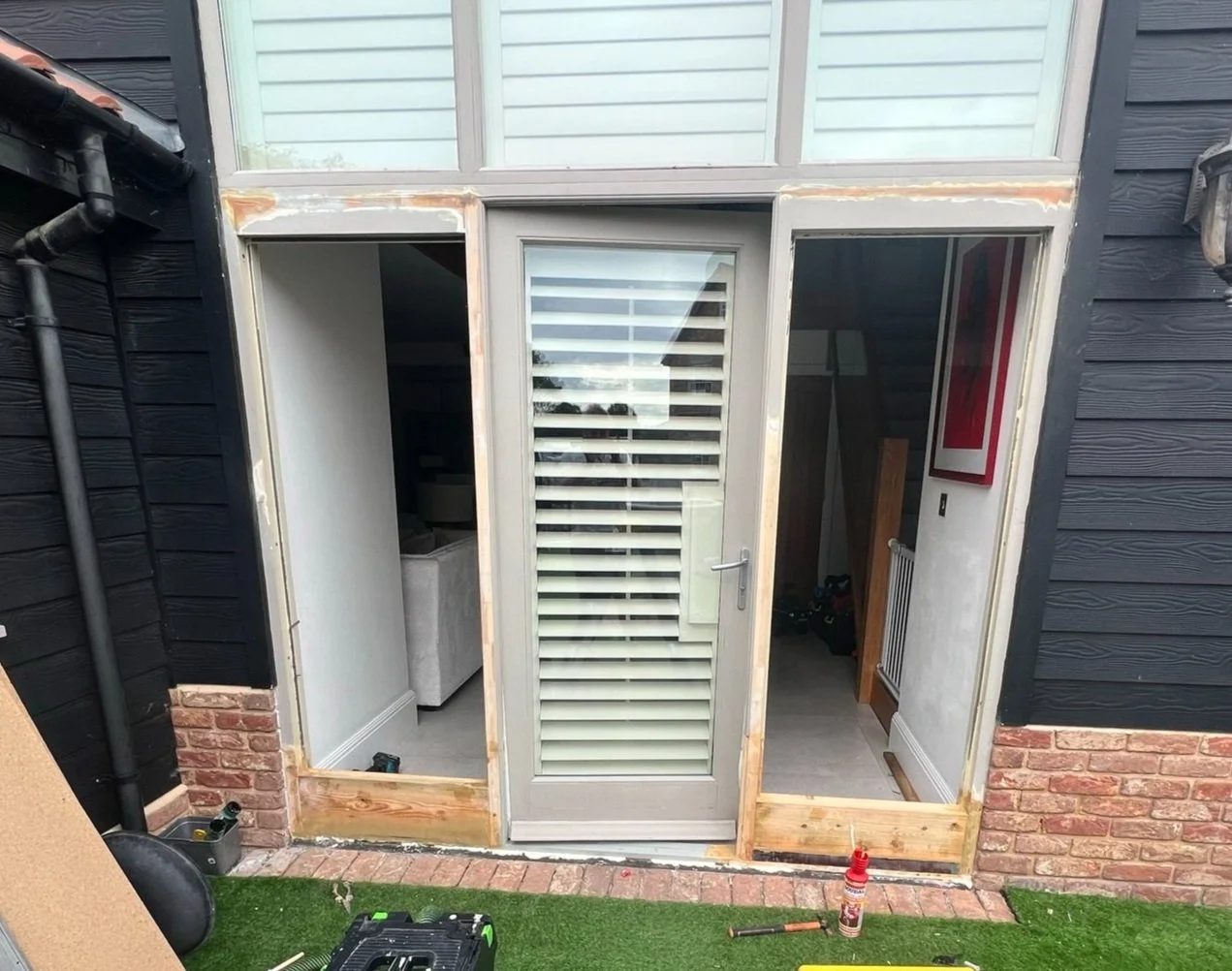 Wooden double door frame under construction with partially installed glass doors and surrounding brick and panel walls. Indoor space visible through the opening. Tools and construction materials on the ground.