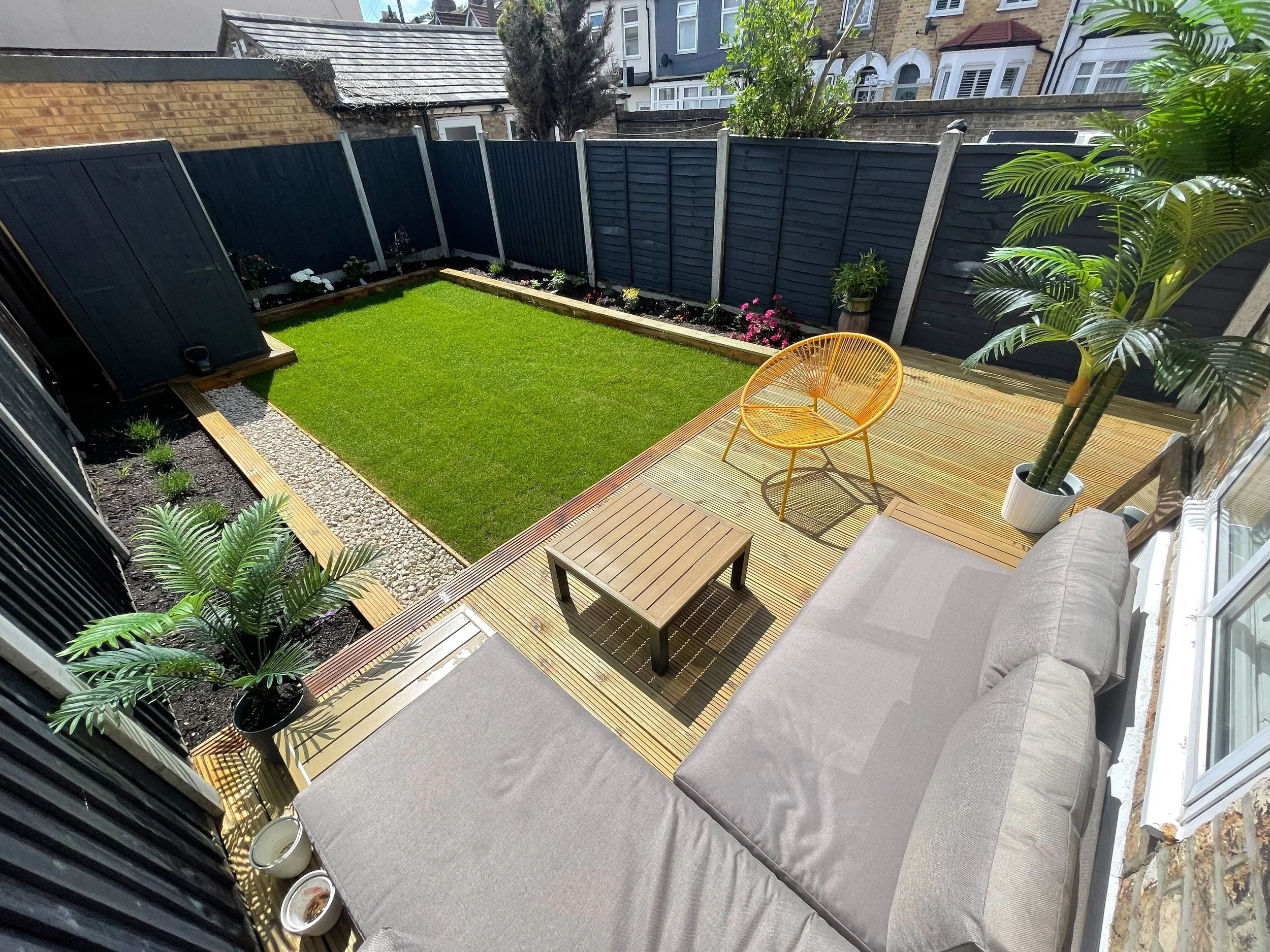 Small backyard with a green lawn, wooden deck, gray L-shaped outdoor sofa, small wooden table, yellow chair, potted plants, and high black fences.