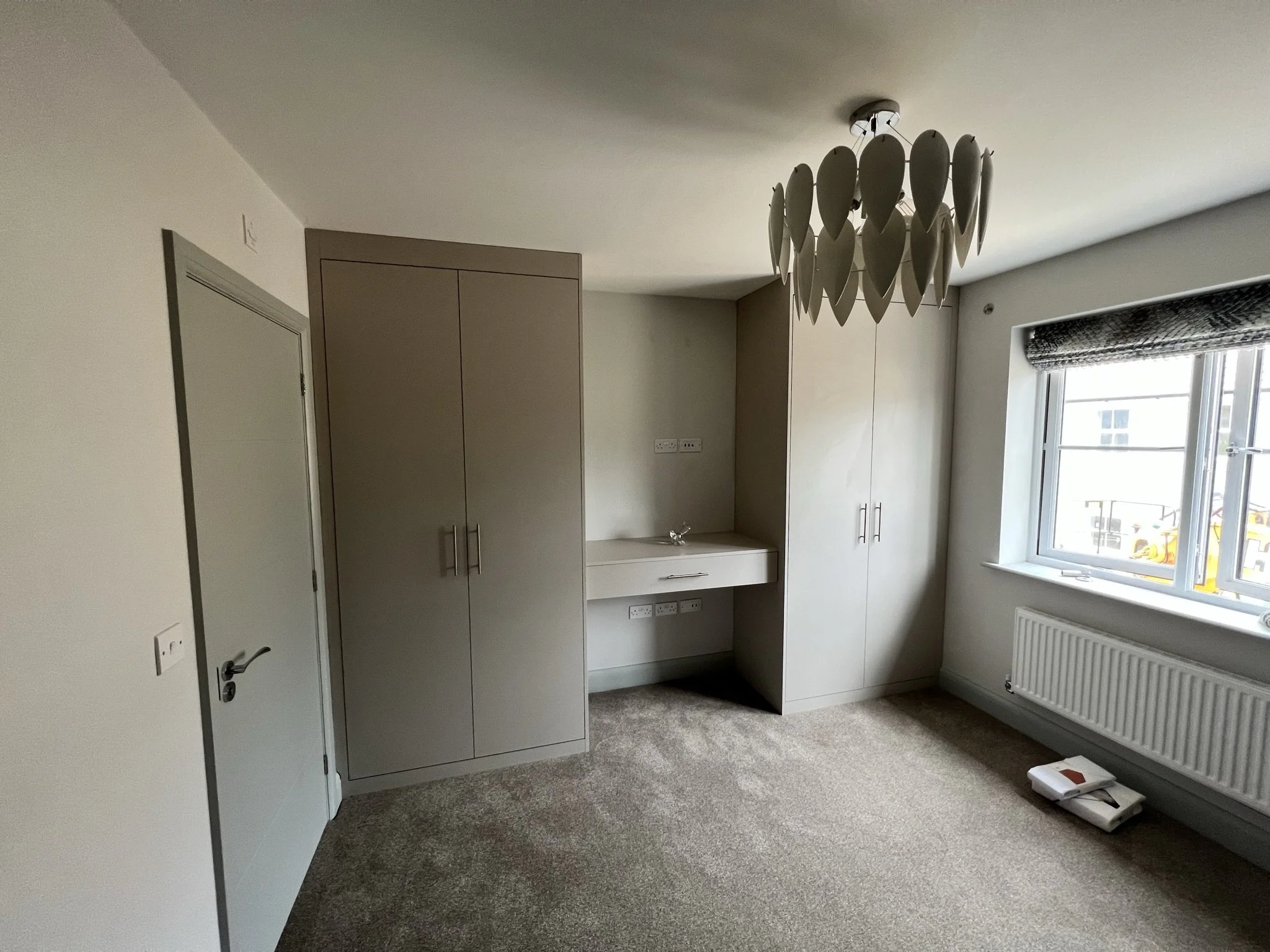 Modern bedroom interior with built-in wardrobes, a unique chandelier, and a window with a partial view. The carpeted floor is light-colored, and there are a few items placed on the floor near the window, including a book and a stack of papers.