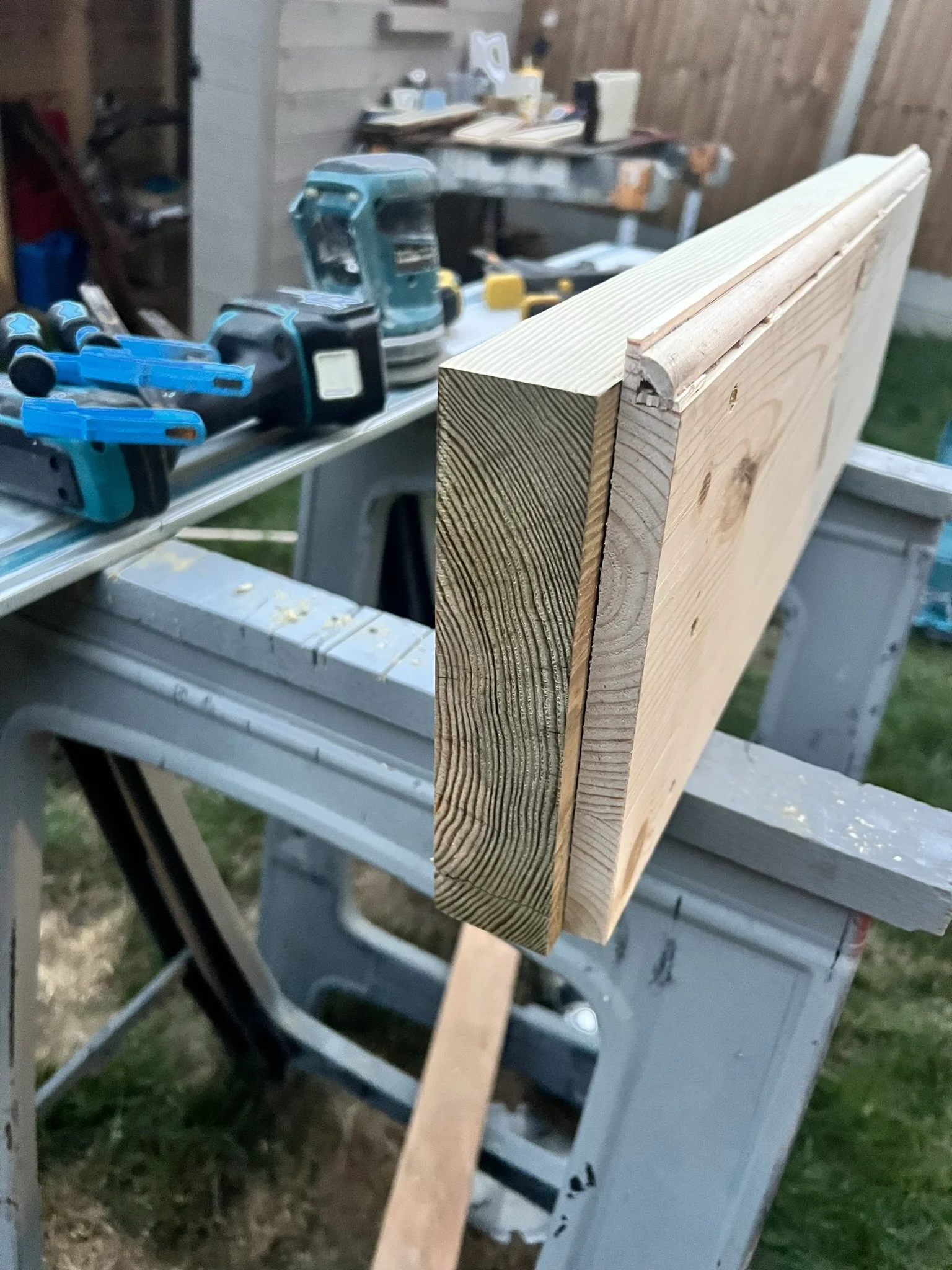 A close-up of a wooden board with a trimmed edge on a sawhorse, surrounded by various carpentry tools including clamps, a planer, and a sander. The background shows a workshop environment with wooden materials.