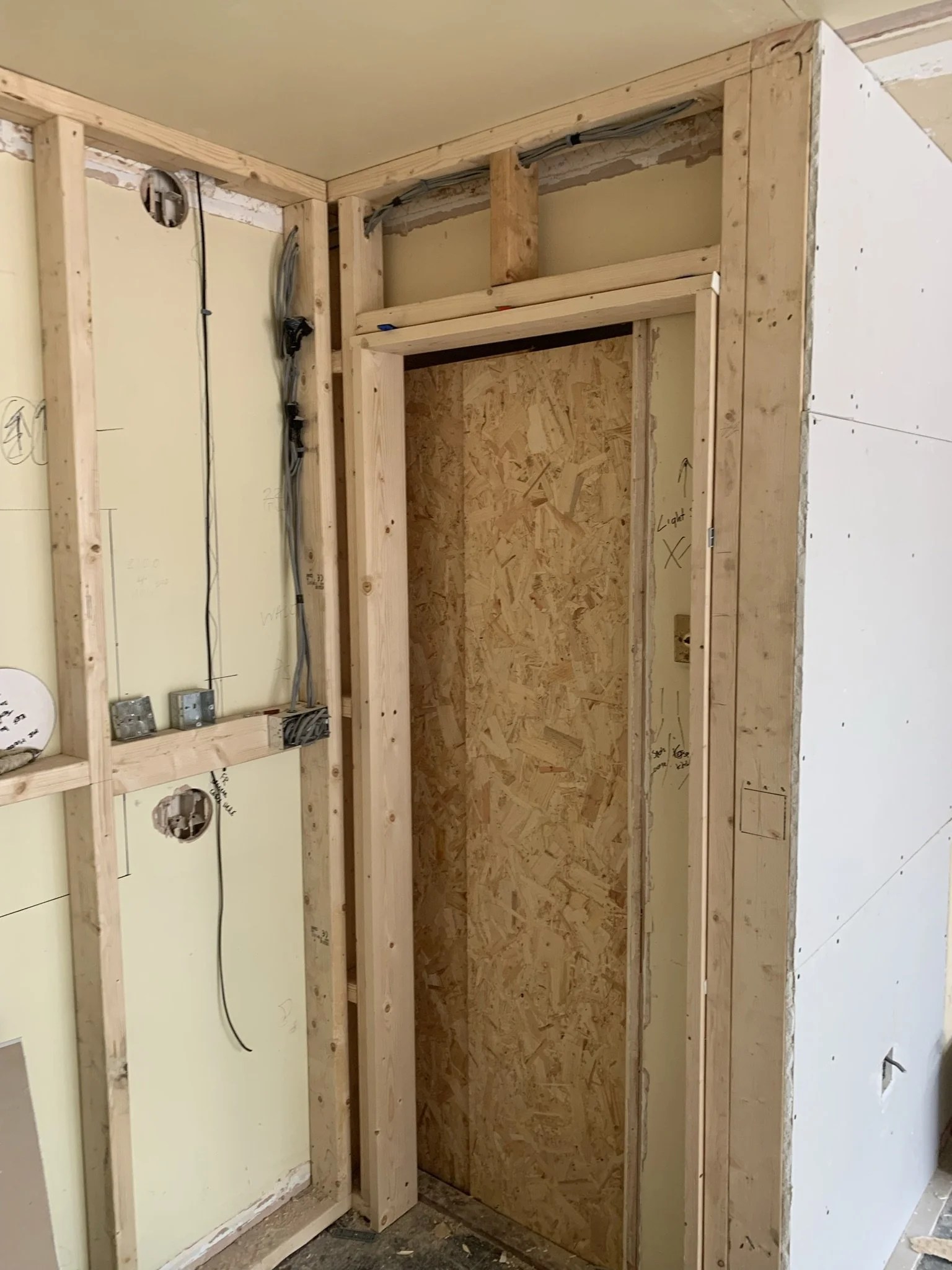 Interior view of a partially constructed room featuring exposed wooden framing, insulation, and electrical wiring. The room has a door partially covered with oriented strand board (OSB) and drywall on the walls.