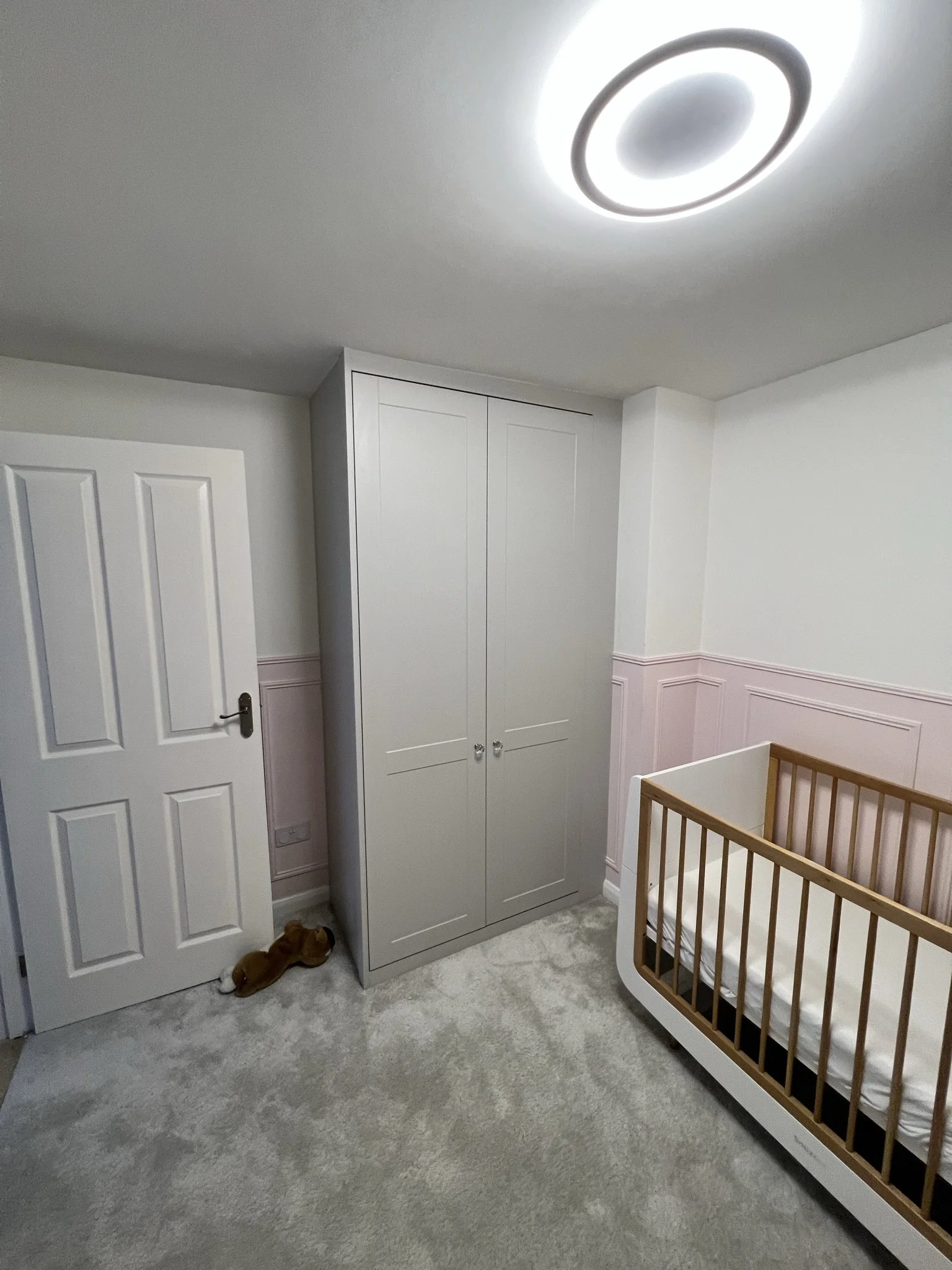 Nursery room with gray wardrobe, wooden crib, white door, plush toy on floor, and circular ceiling light.