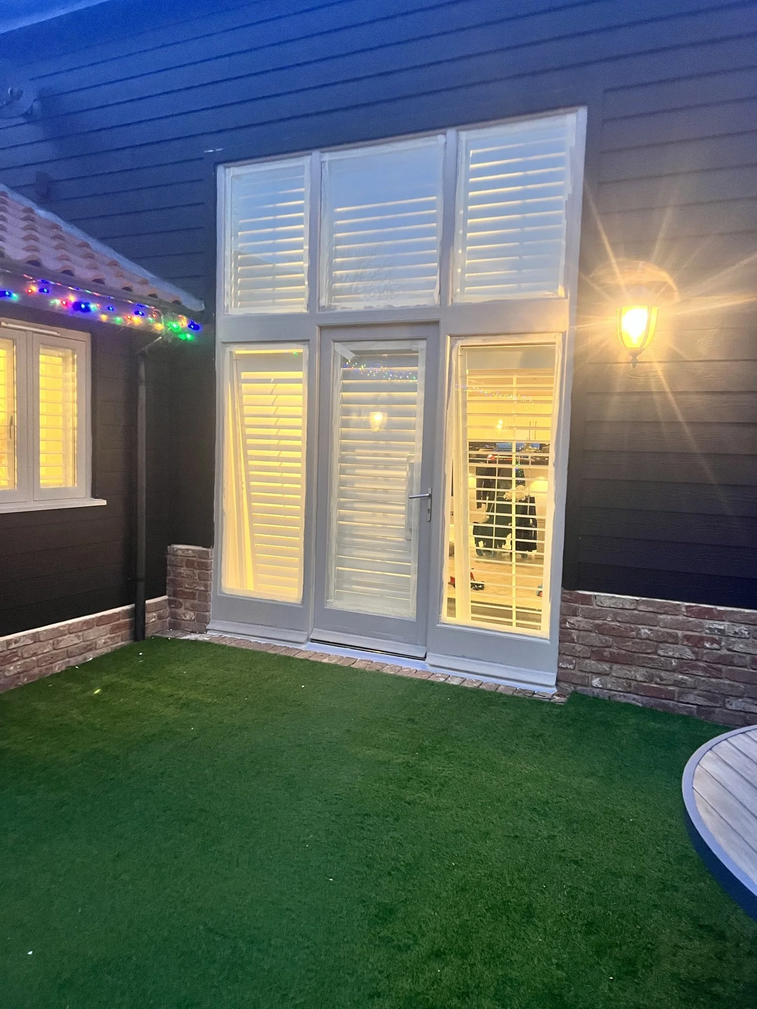 Exterior view of a building with a door, tall windows, white shutters, and a wall light. Holiday string lights are visible on the roof edge. The ground is covered with artificial grass.