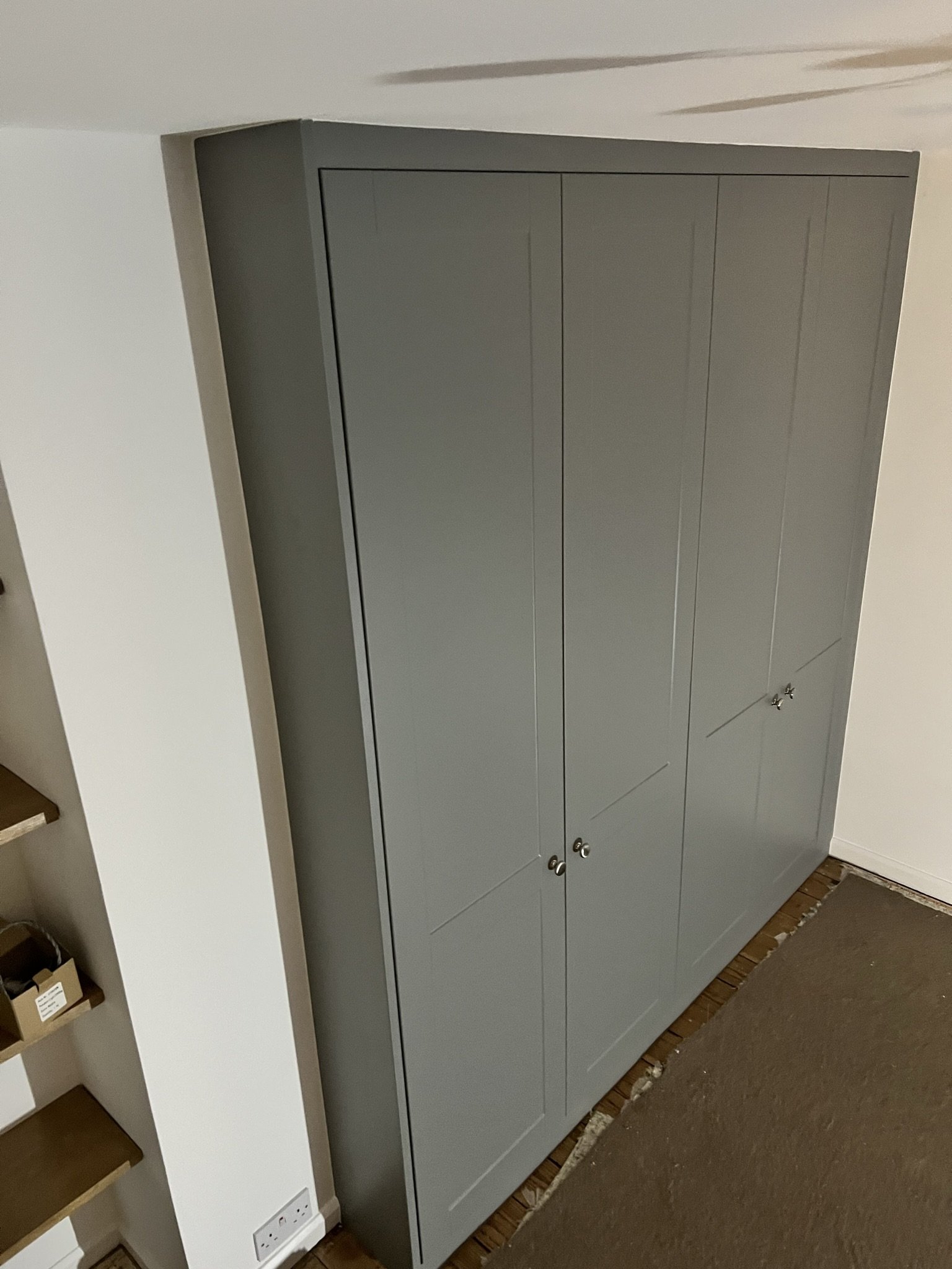 Gray wardrobe with three doors in a room corner, adjacent to shelving with a box, and an electrical outlet visible nearby.