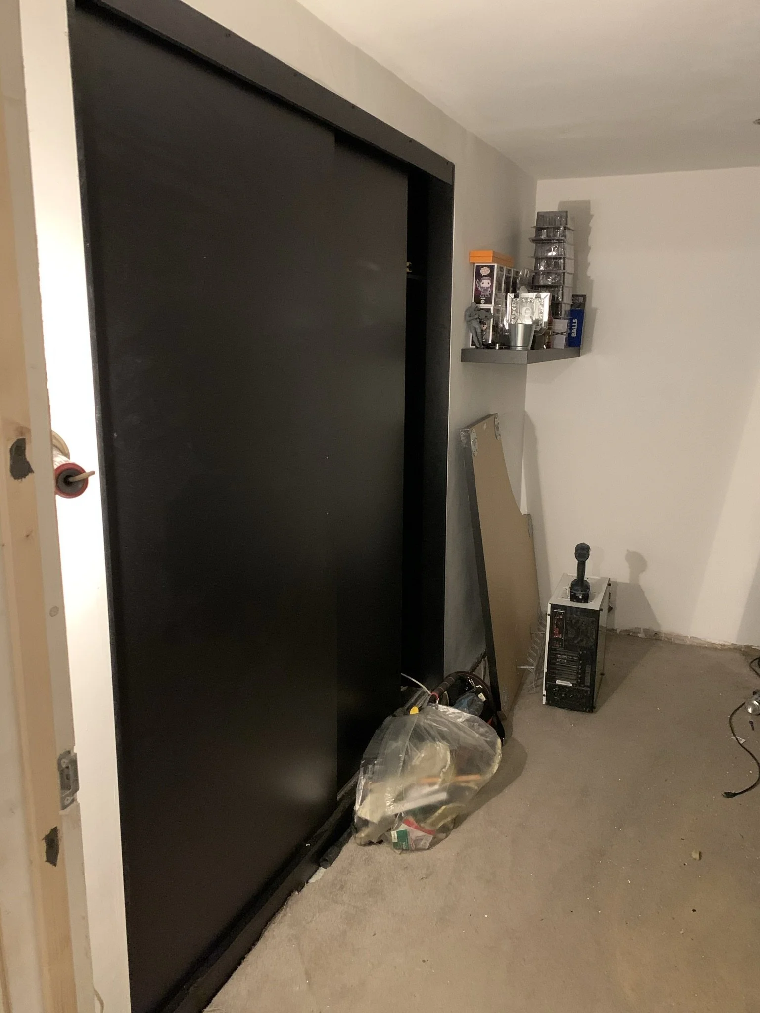 Room with a black sliding door, a transparent bag filled with miscellaneous items on the floor, a metal shelf with various objects, and an open computer case on the ground.