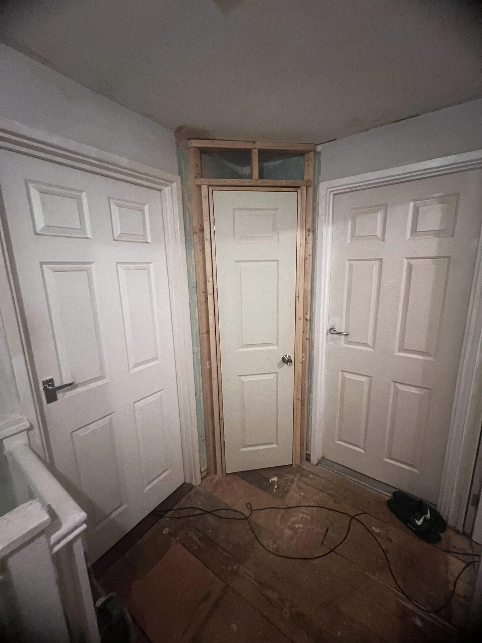 Three white interior doors in a dimly lit hallway, with a wooden frame surrounding the center door. The floor is unfinished wood with cables strewn across.