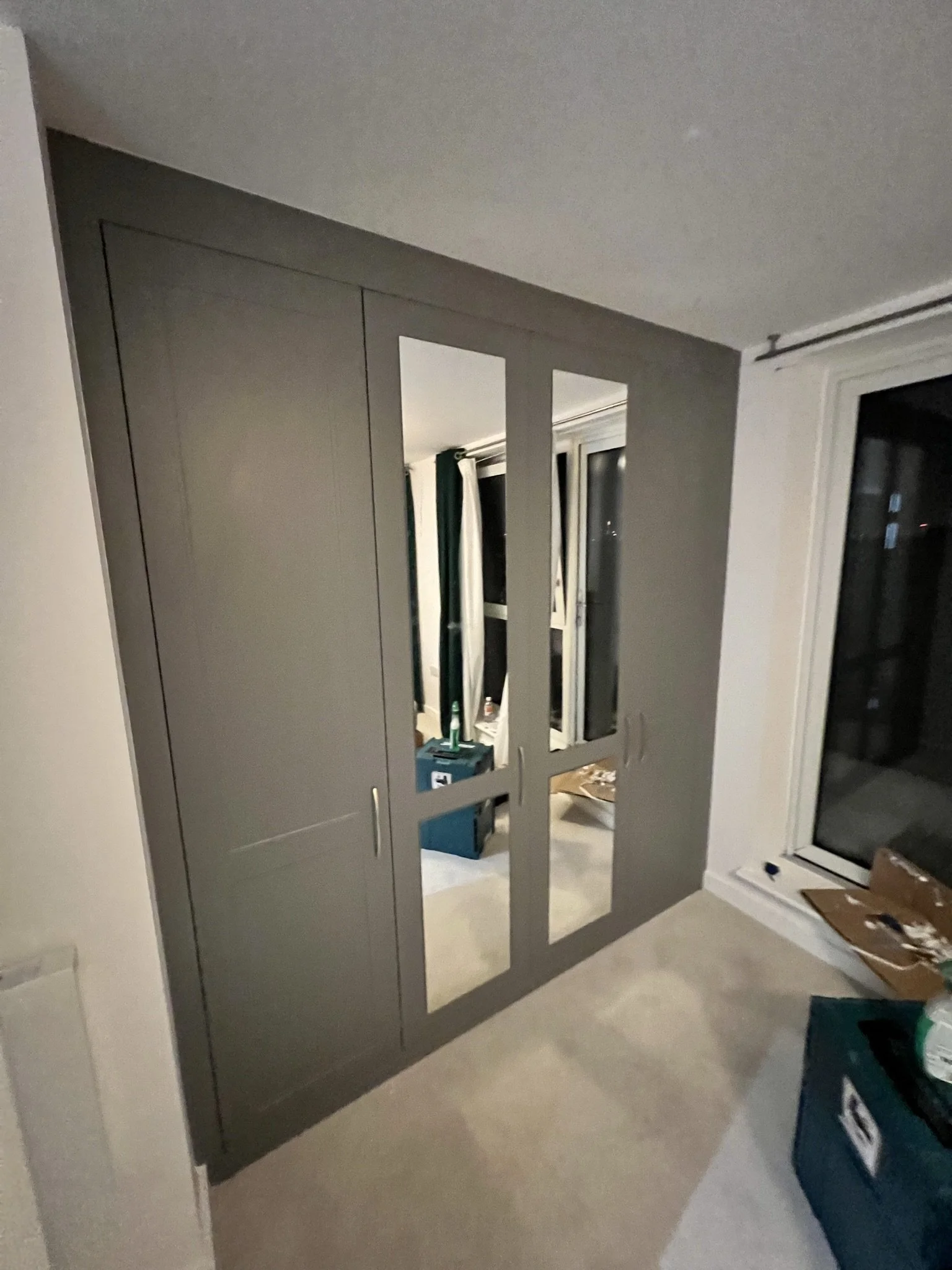 Gray built-in wardrobe with mirrored doors in a carpeted room, next to large windows.