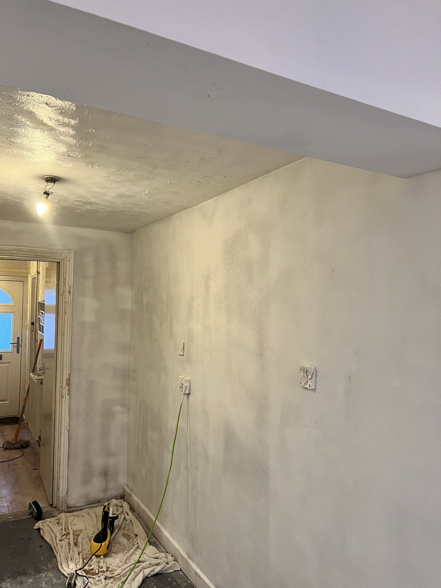 Interior of a room under renovation with white painted walls, a bare light bulb hanging from the ceiling, a door slightly open, and some tools like a sander and a power cord on a sheet-covered floor.
