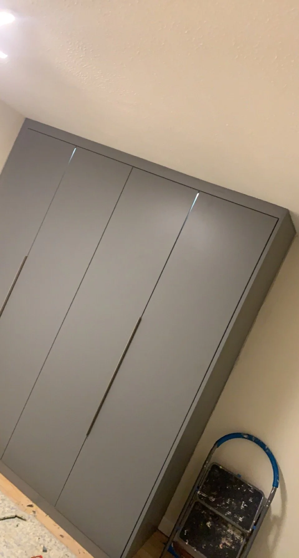 Gray wardrobe with closed doors next to a folded step ladder in a room.