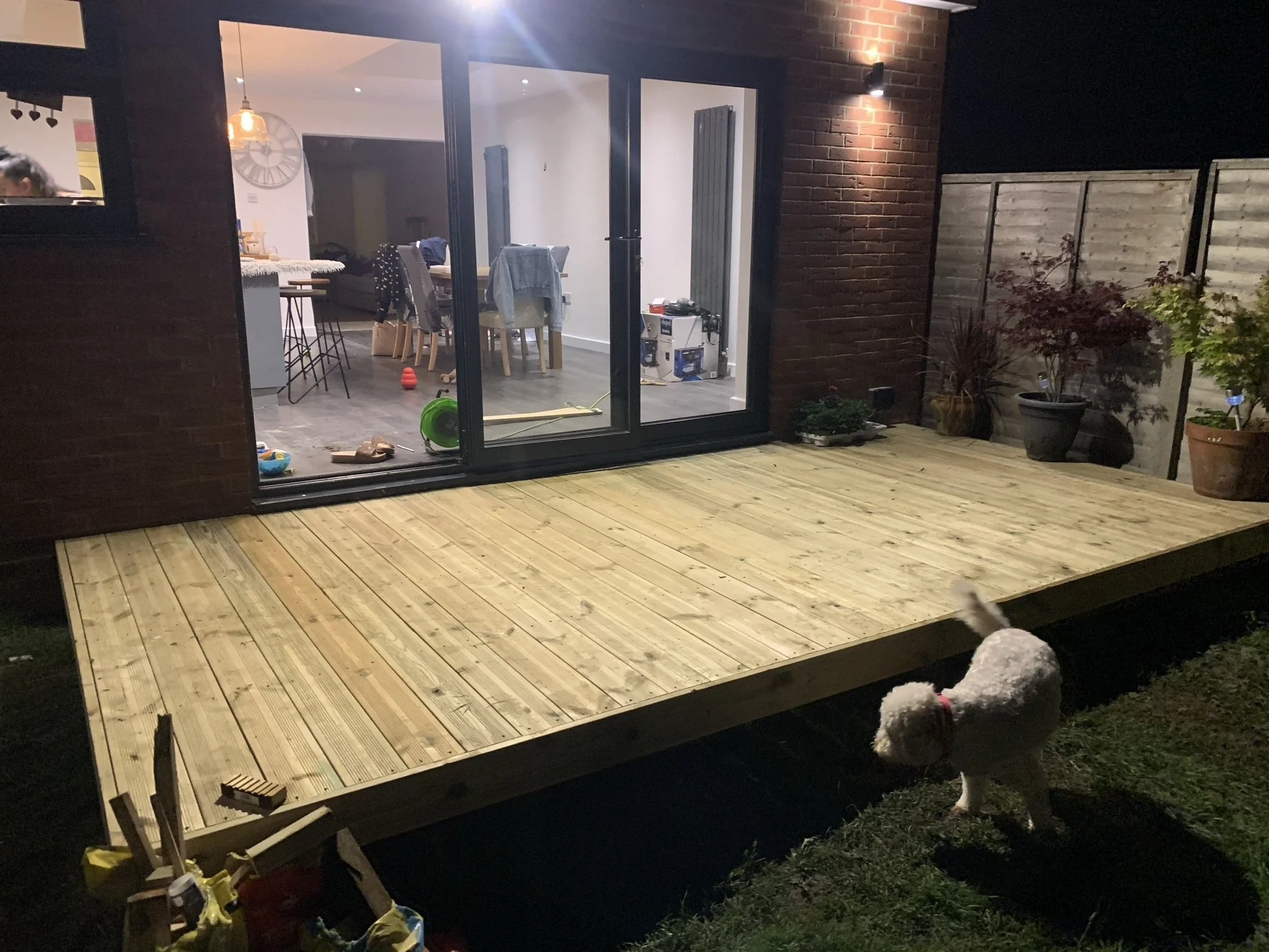 A newly built wooden deck attached to a house with sliding glass doors, illuminated by an exterior light at night. Inside the house, a dining area with a hanging light fixture is visible. A white dog is in the foreground on the grass, near the deck.