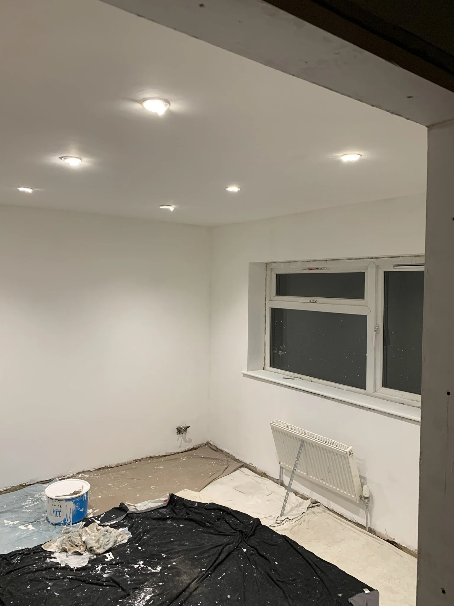 Room under renovation with white painted walls, recessed ceiling lights, a window, and a radiator. Floor covered in protective sheets, and paint supplies visible.