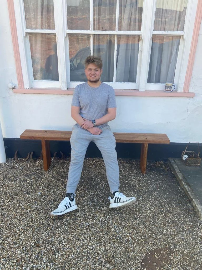 A person with brown hair sits on a wooden bench outside in front of a window. They are wearing a light gray t-shirt, gray sweatpants, and white sneakers. There is a mug on the windowsill.