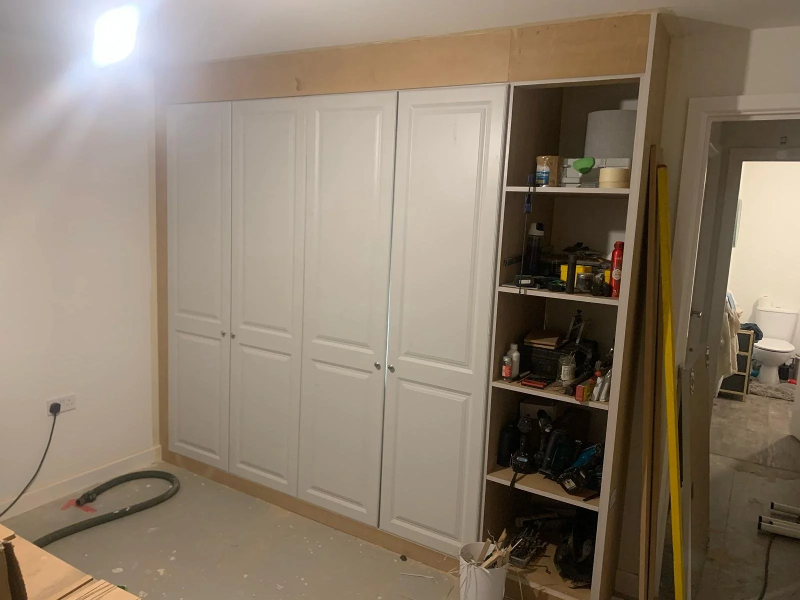 White built-in wardrobe with closed doors and adjacent open shelving filled with tools and household items. A bathroom is visible in the background.