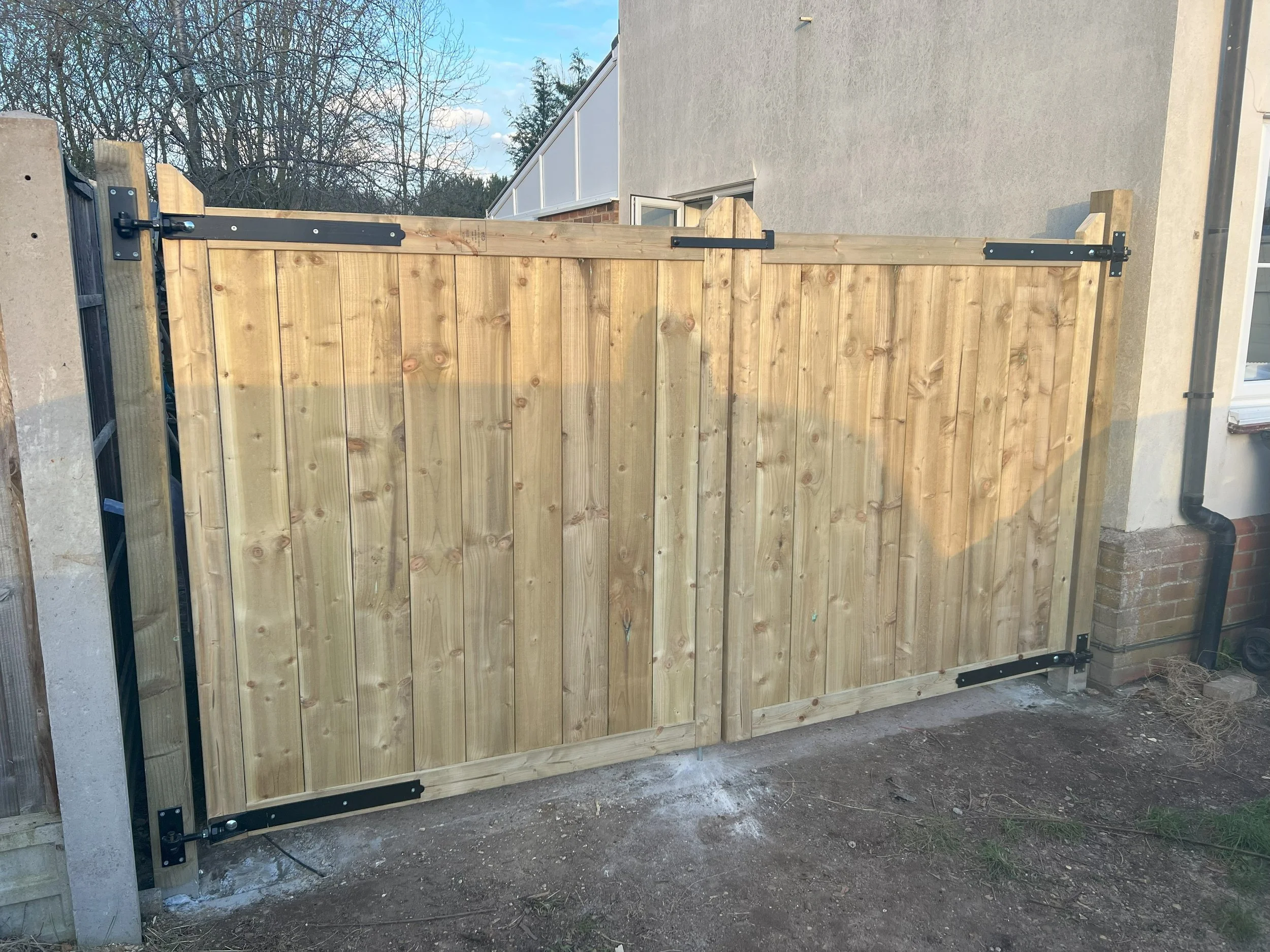 Wooden double gate with black metal hinges and latches, set in a backyard with bare ground.