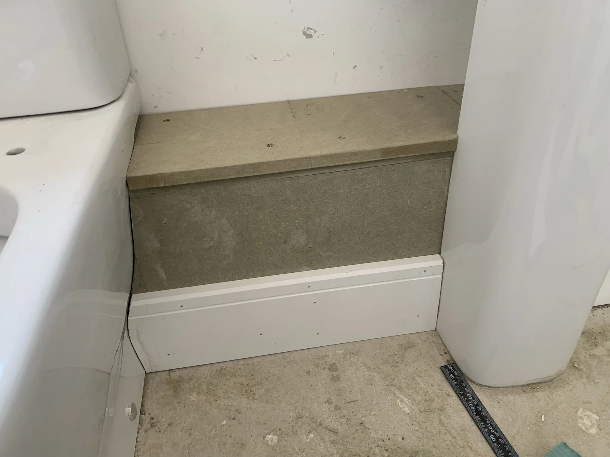 A beige step or ledge in a room corner positioned between a bathtub and a white pedestal or pillar. The floor is unfinished concrete and a blue measuring tool is on the ground.