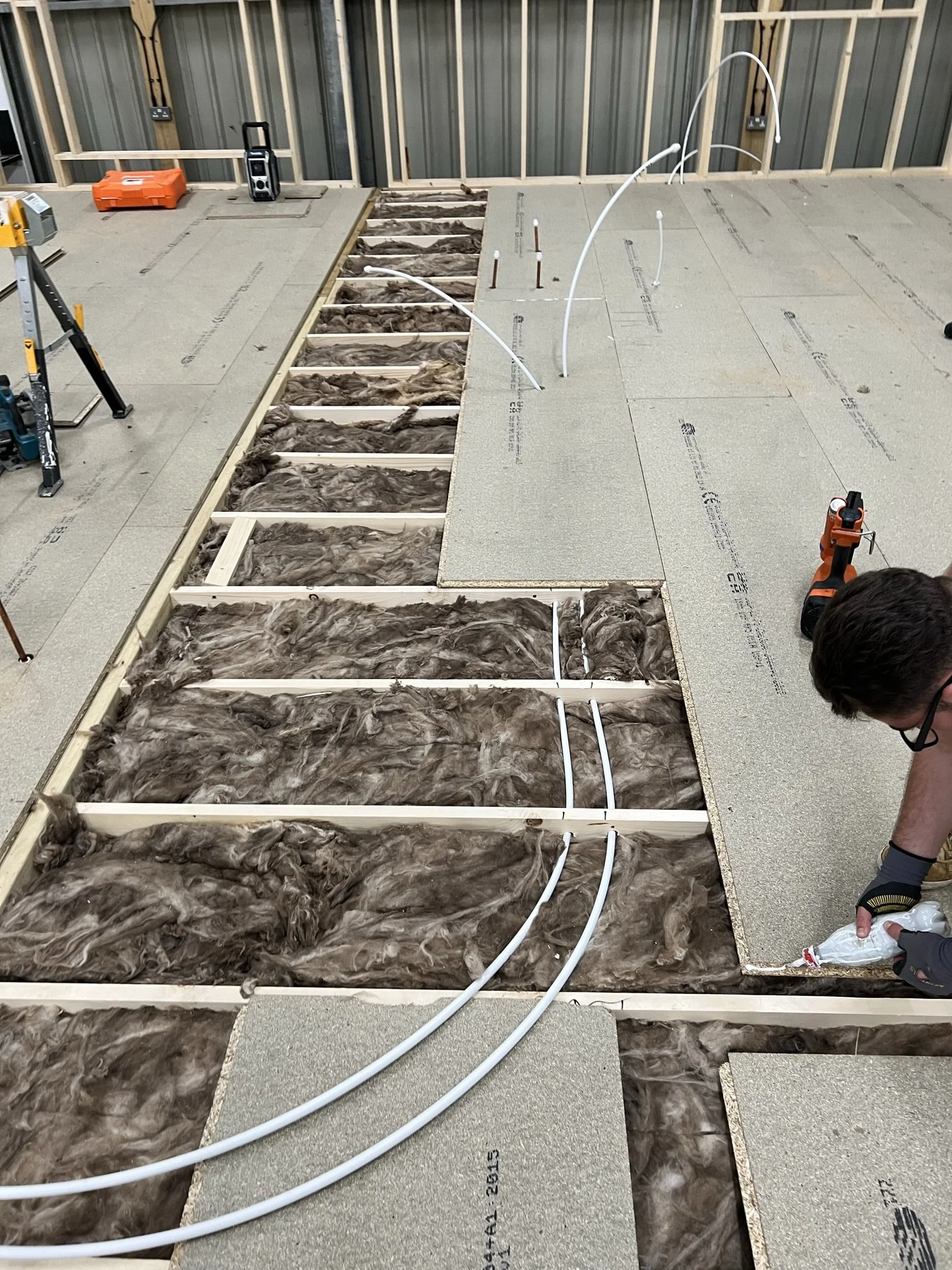 Construction site with floor insulation and wooden framework, white pipes being installed, person applying adhesive, and power tools visible.