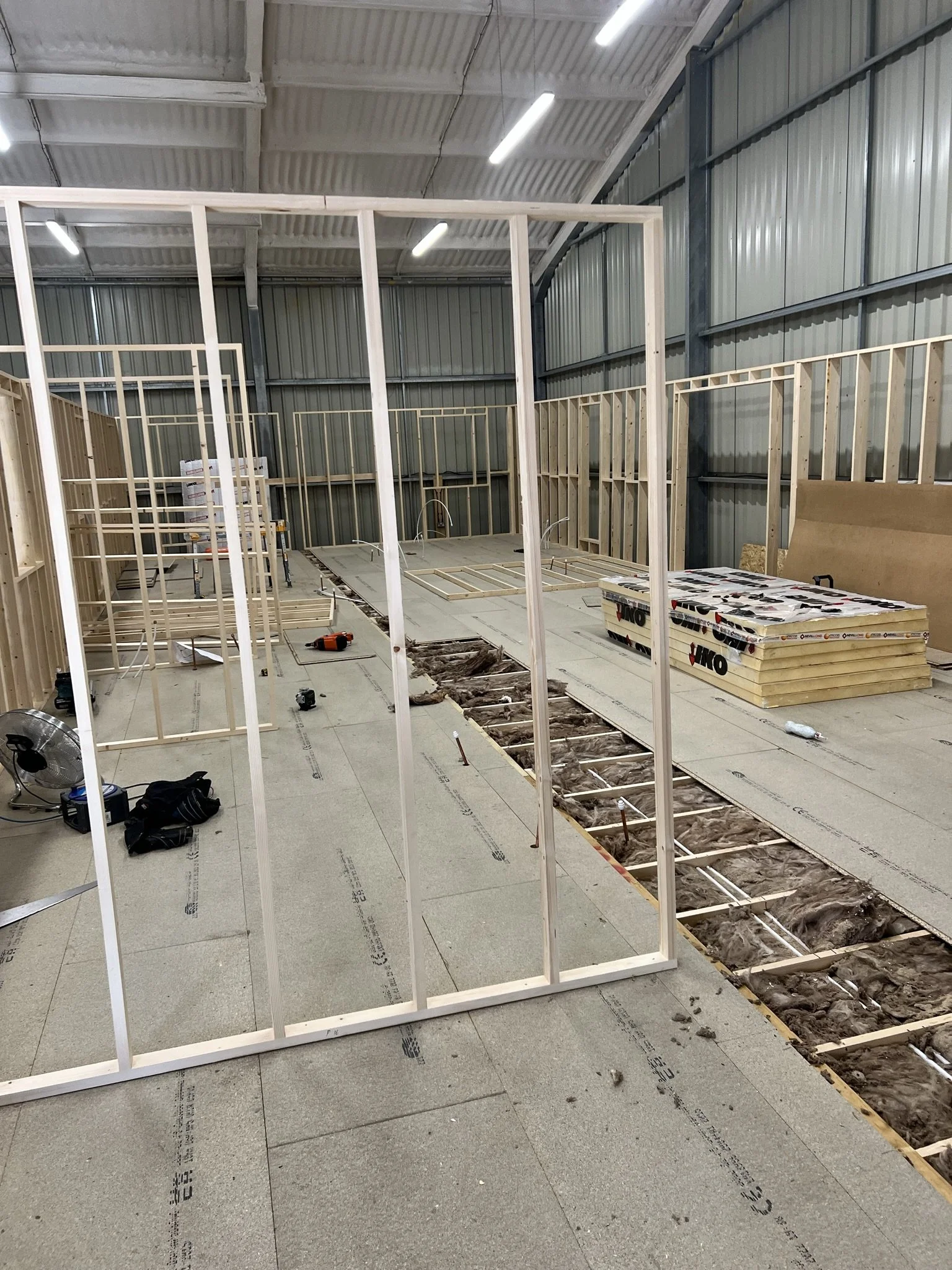 Interior construction site with framing structures, exposed floor insulation, and building materials.