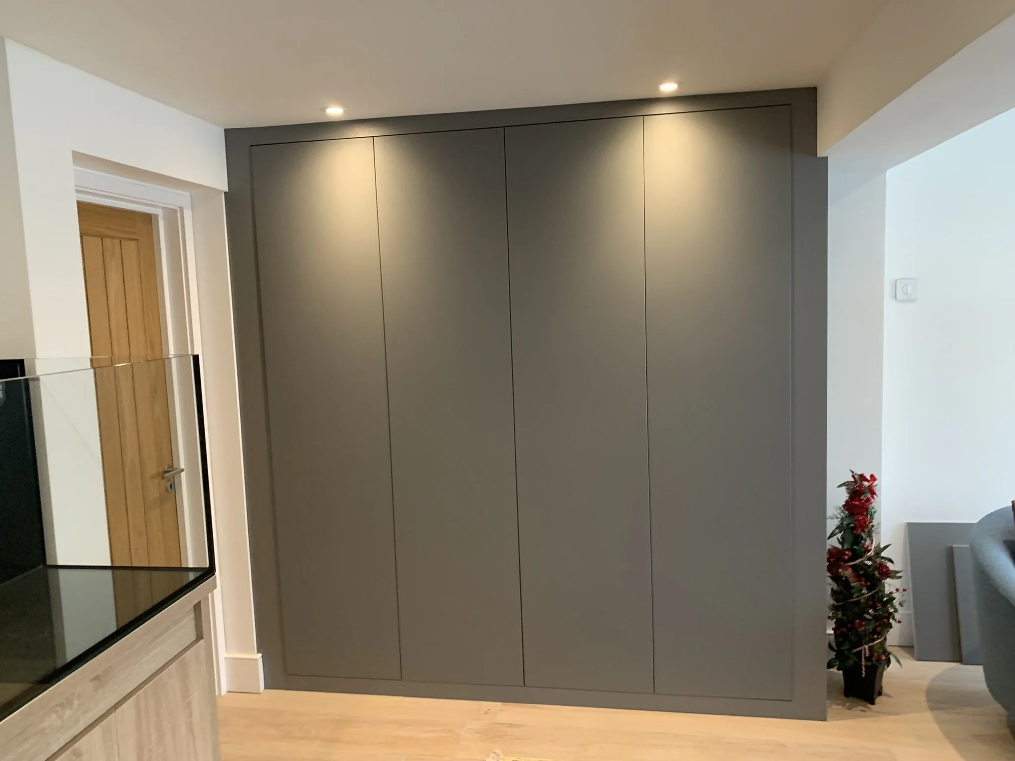 Modern, gray wall-mounted cabinet with three panels, two overhead spotlights, and adjacent wooden door, in a room with light-colored flooring and a small decorative plant on the side.