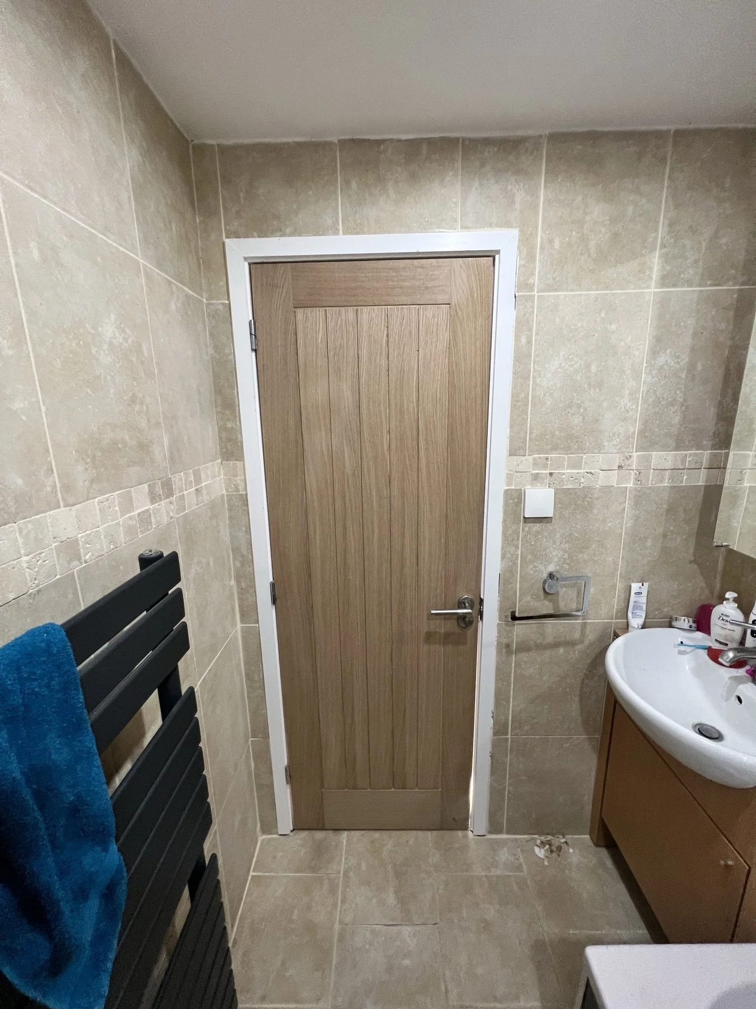 Bathroom interior with a wooden door, beige tiled walls, a black towel rack with a blue towel, a white sink with toiletries, and a wooden cabinet.