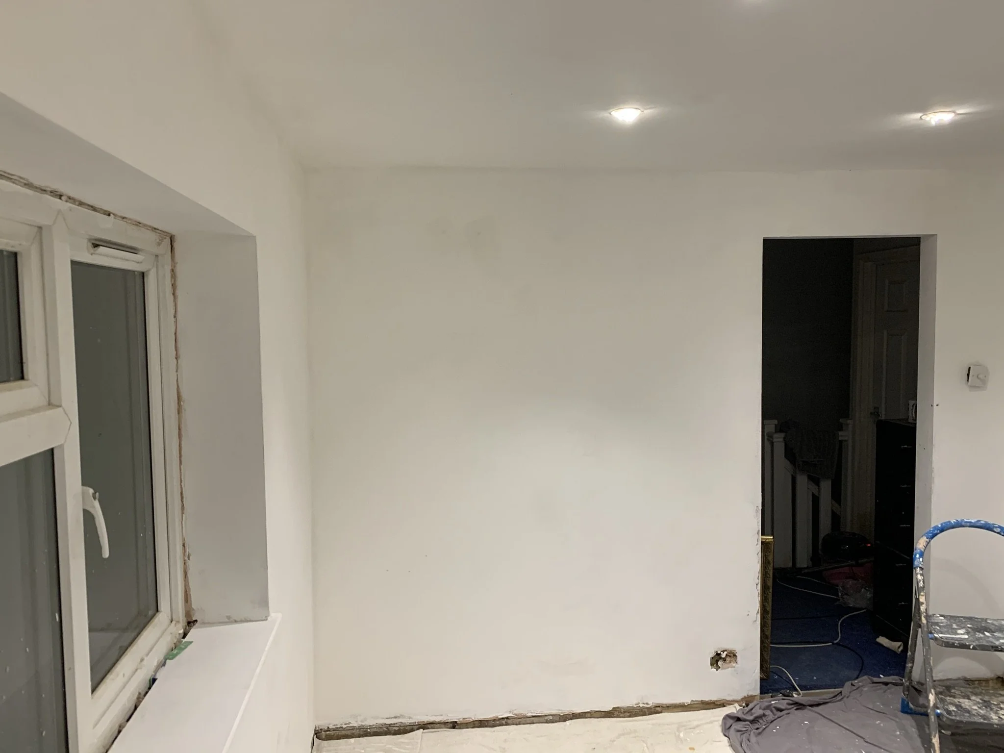 Interior room under renovation with white painted walls, window, recessed lights, ladder, and open doorway.
