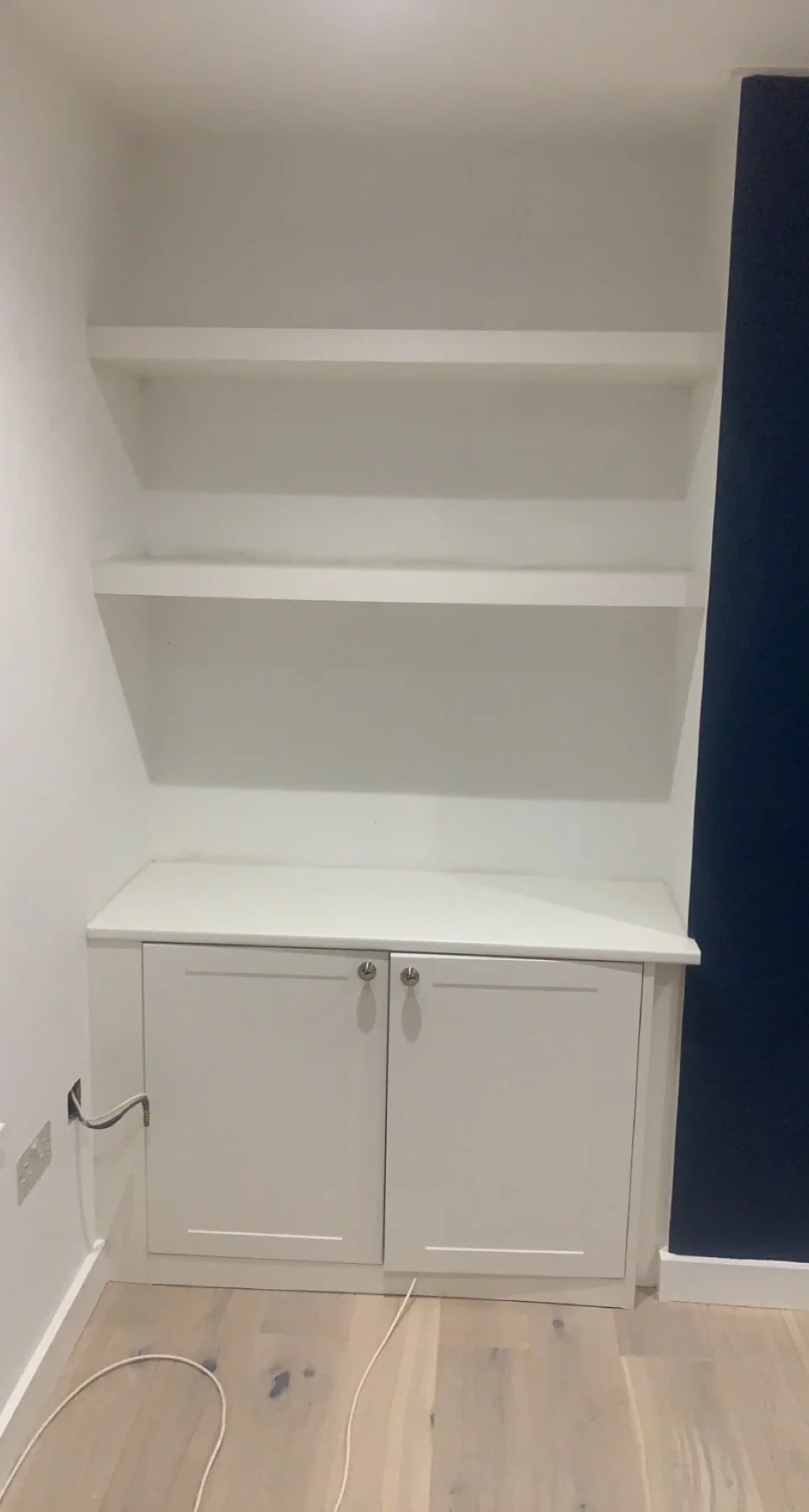 White built-in shelves and cabinet with two closed doors, white walls, and light wood flooring.