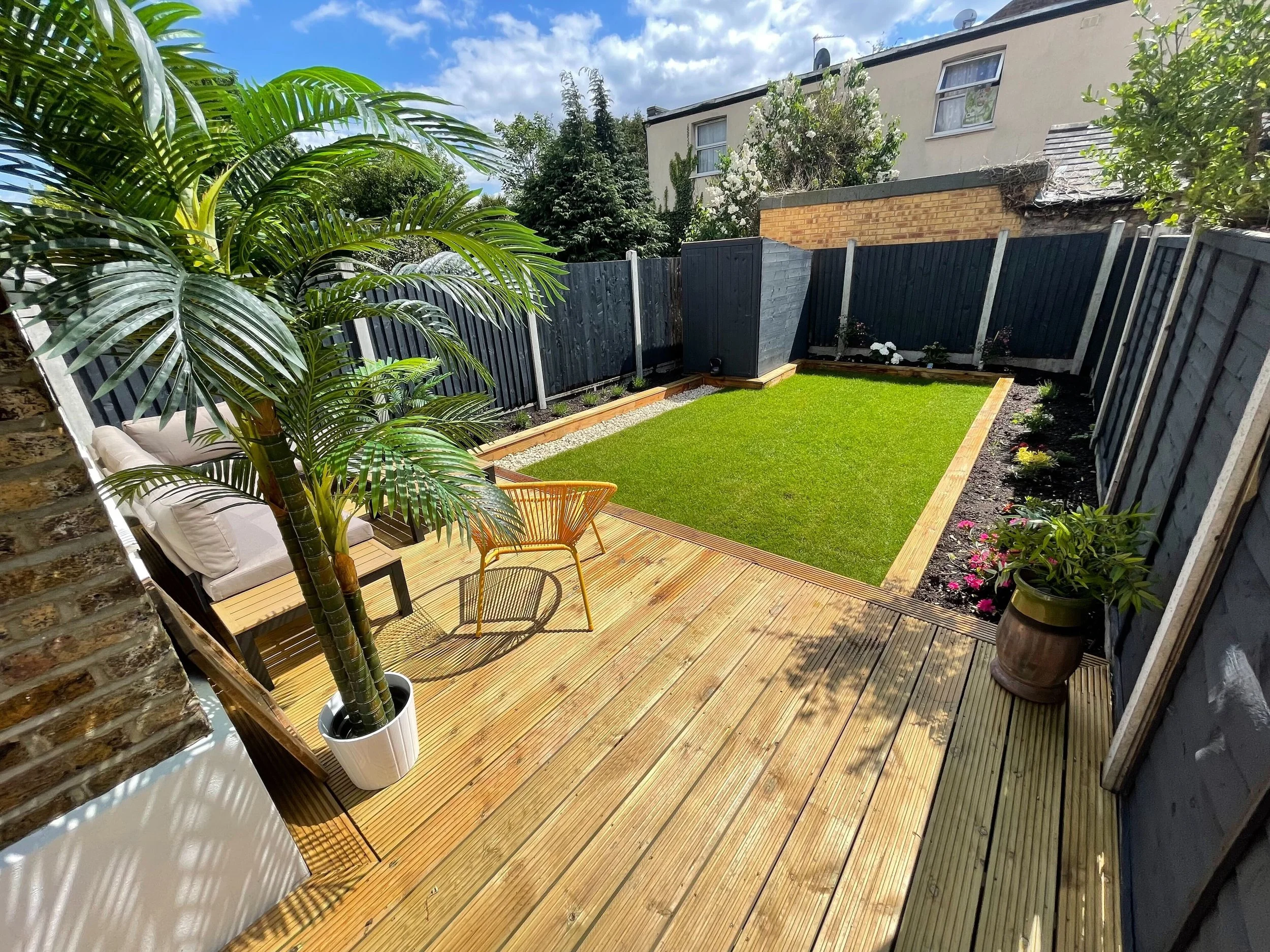 Backyard with wooden deck, potted palm tree, seating area, and small grass lawn surrounded by fence.