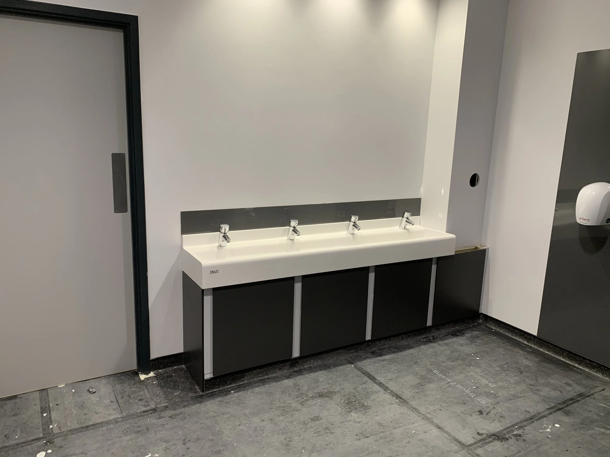 Public restroom with a triple sink and faucets, gray walls, and a hand dryer on the right side. The floor is dark and the restroom door is on the left.