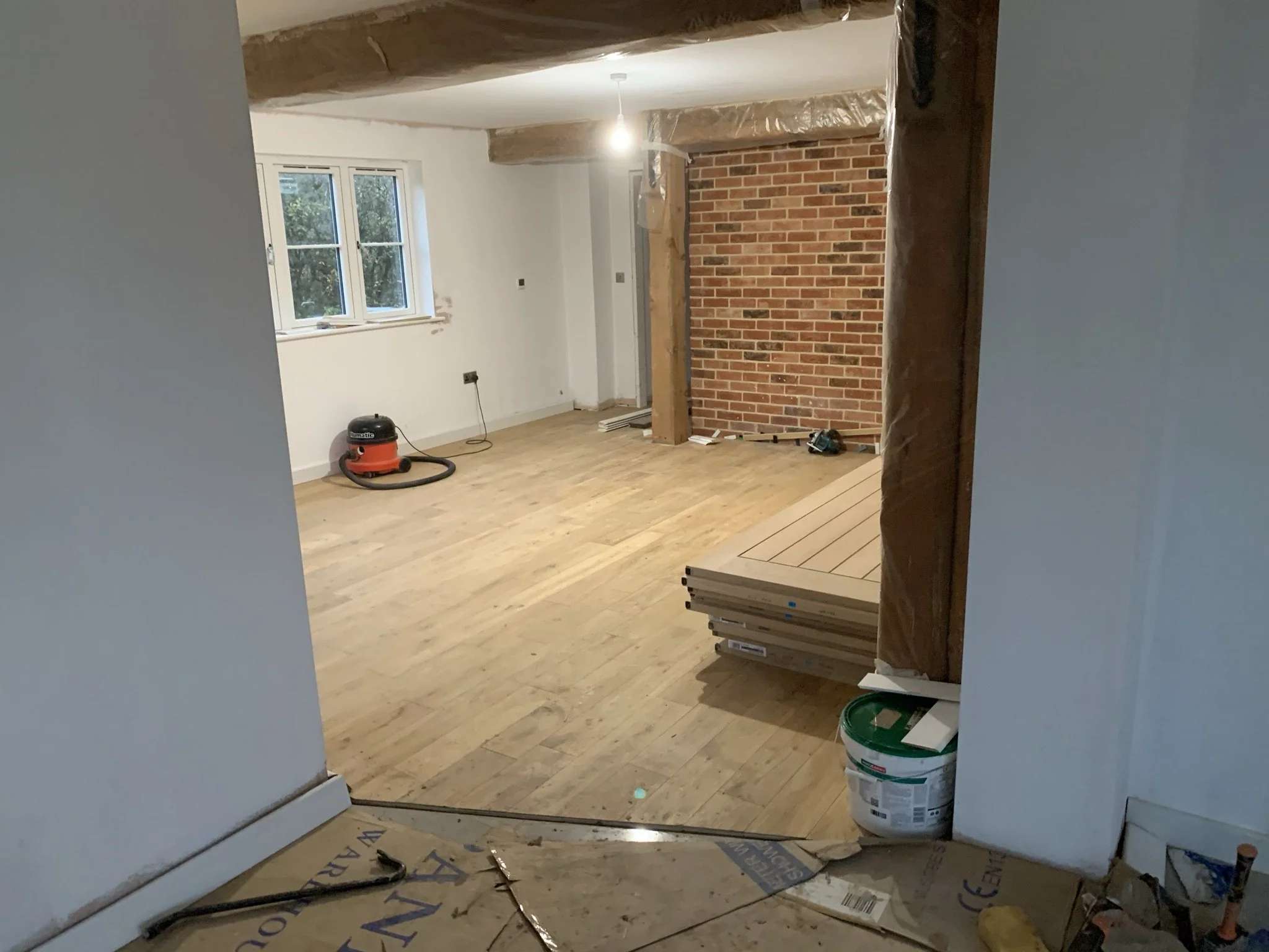 Room under renovation with exposed brick wall, wooden floors, unfinished walls, a stack of wooden planks, paint can, vacuum cleaner, and construction materials.