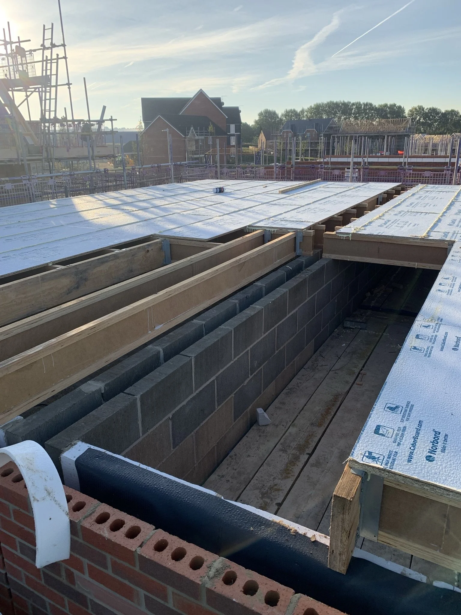 A construction site with exposed brickwork, wood beams, and insulation panels. In the background, there are scaffoldings and partially constructed buildings under the blue sky.