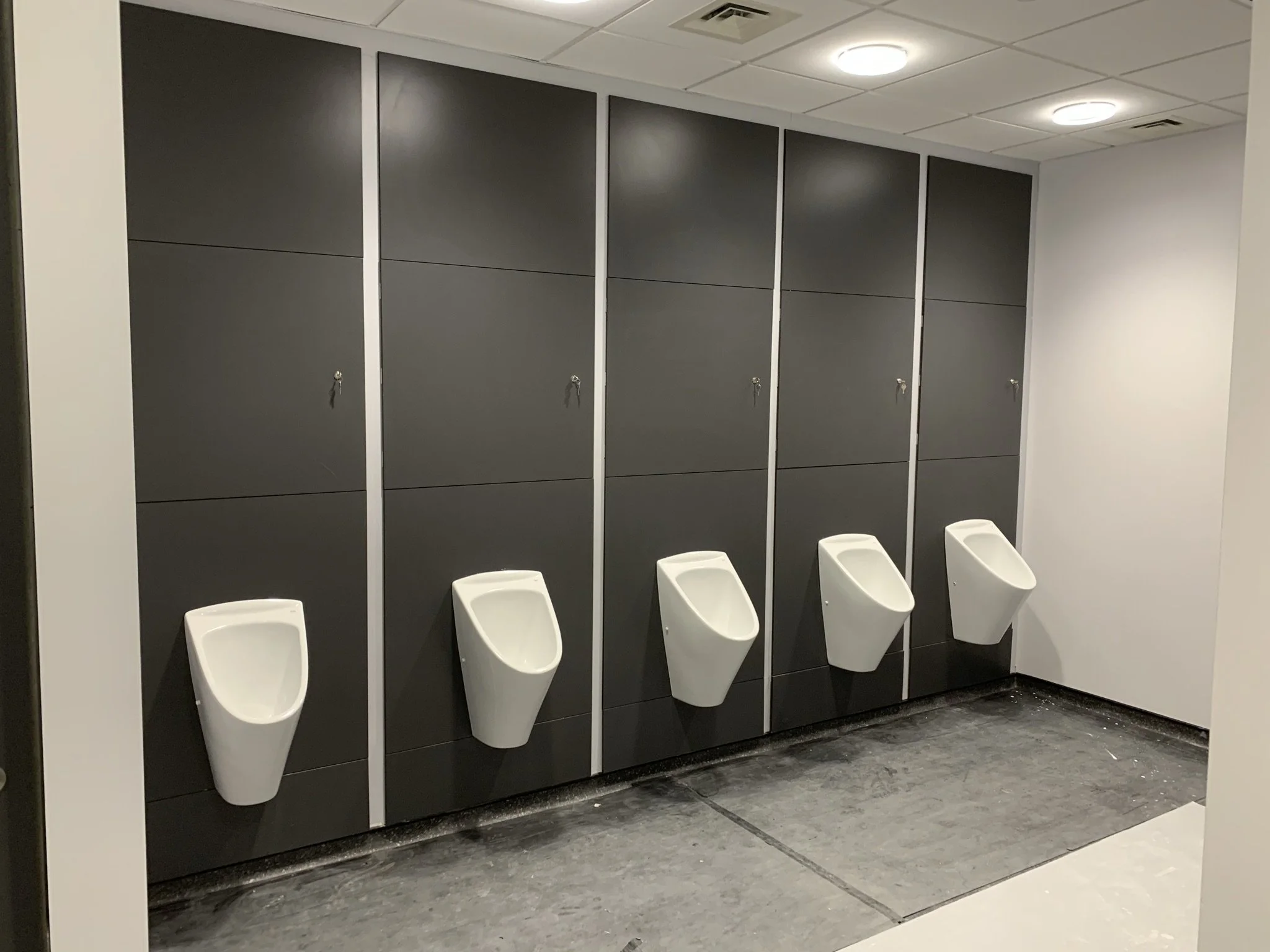 Row of five wall-mounted urinals in a public restroom with dark gray wall panels and white flooring.