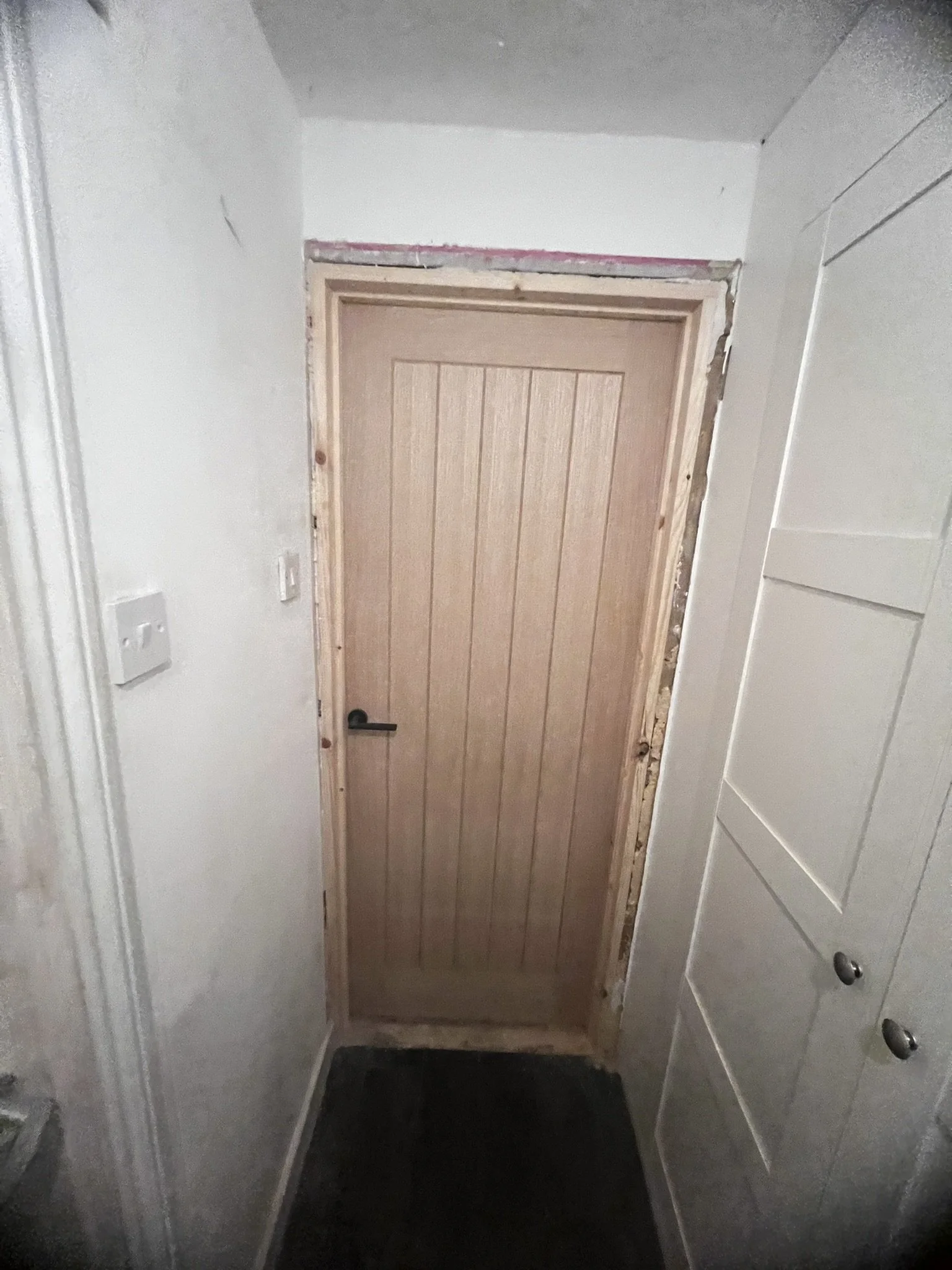 Interior view of a wooden door with a black handle, set in a white wall. The doorframe appears unfinished and there is a light switch on the left side of the wall.