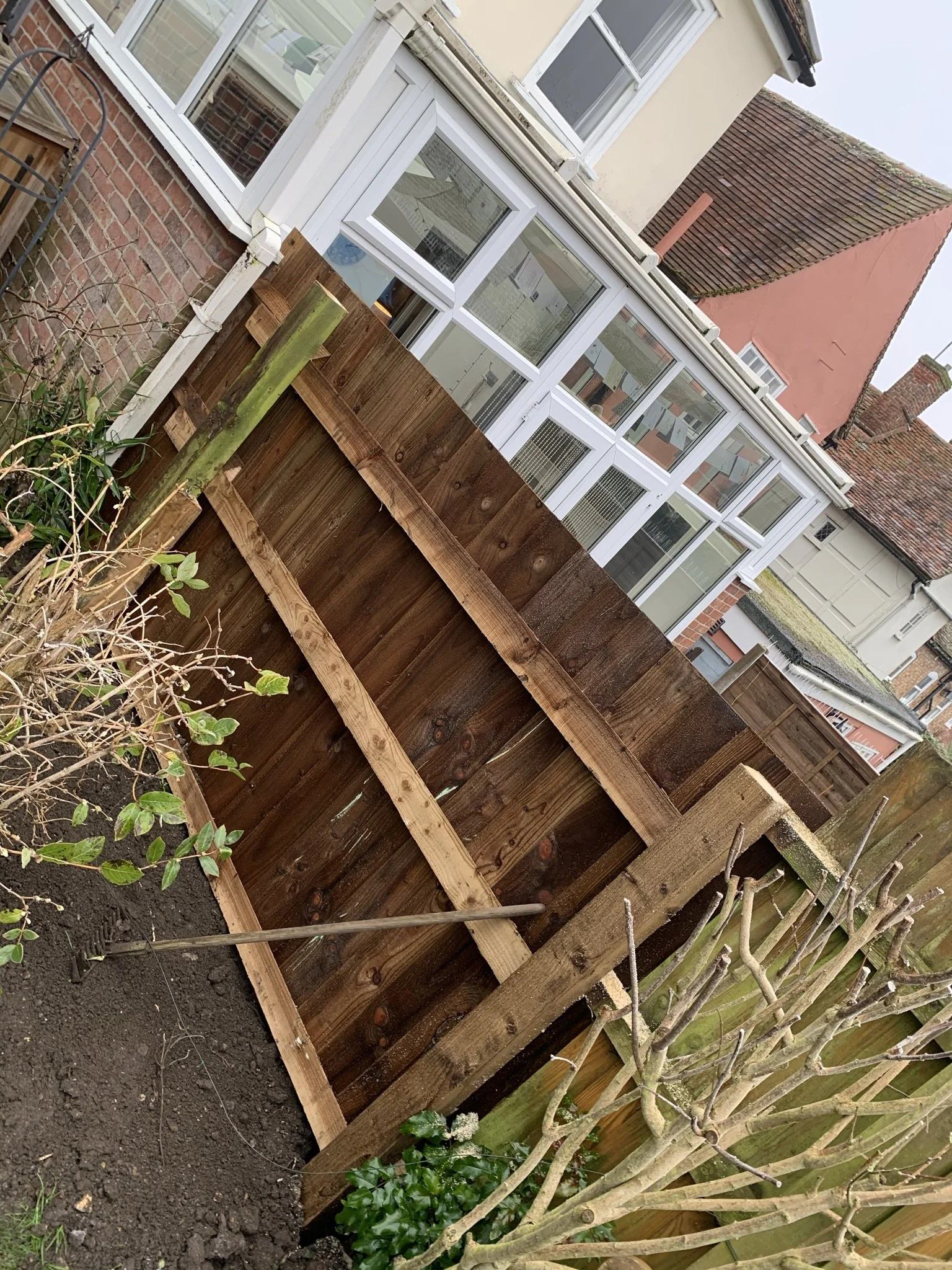 Wooden garden fence under construction near a brick house with windows, a rake leaning against it, and plants nearby.
