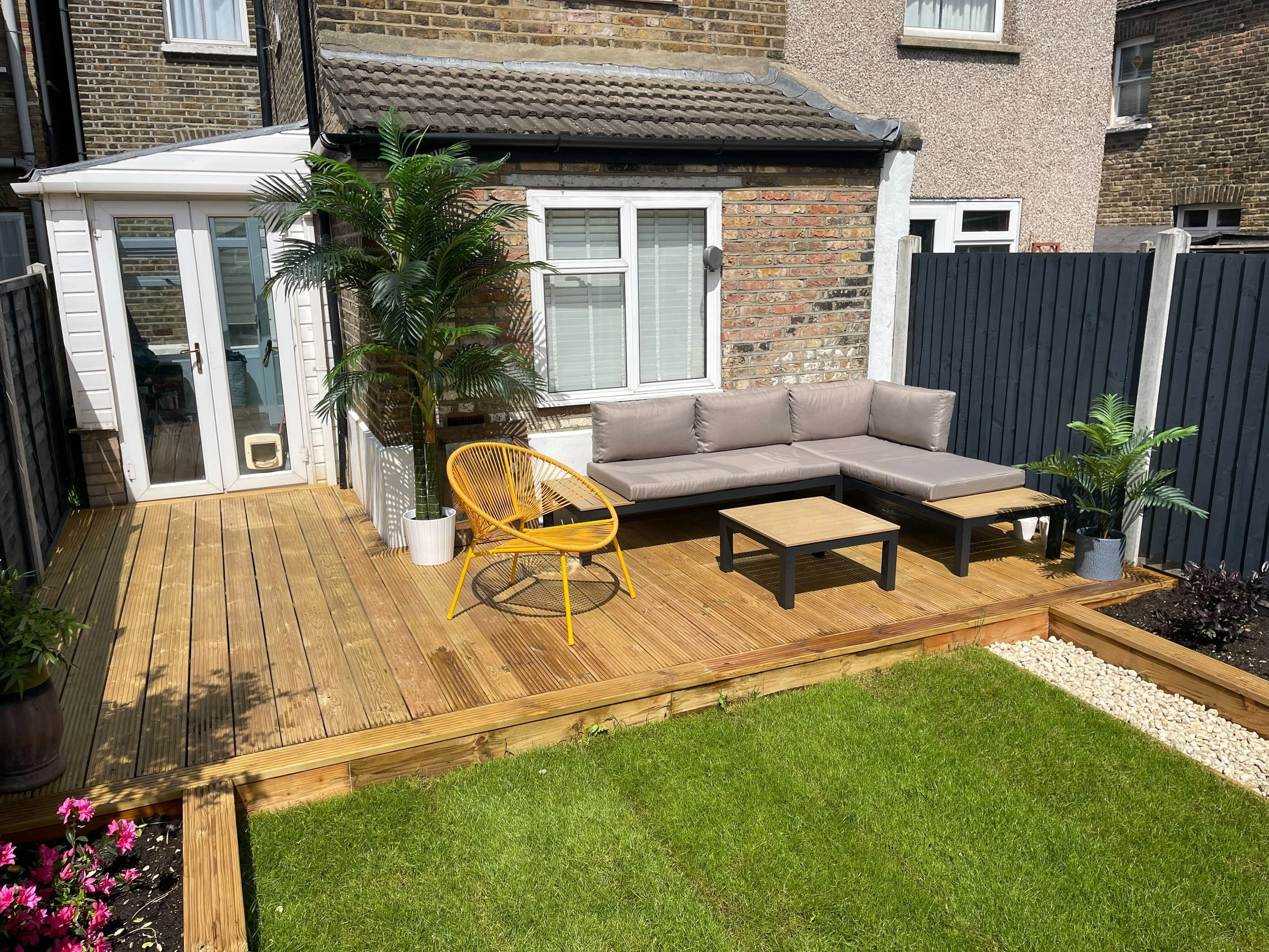 A backyard wooden deck with L-shaped gray patio sofa, a small coffee table, a yellow chair, and potted plants. A brick house with a glass door and a dog door is in the background. Well-maintained lawn and flower bed are visible around the deck.